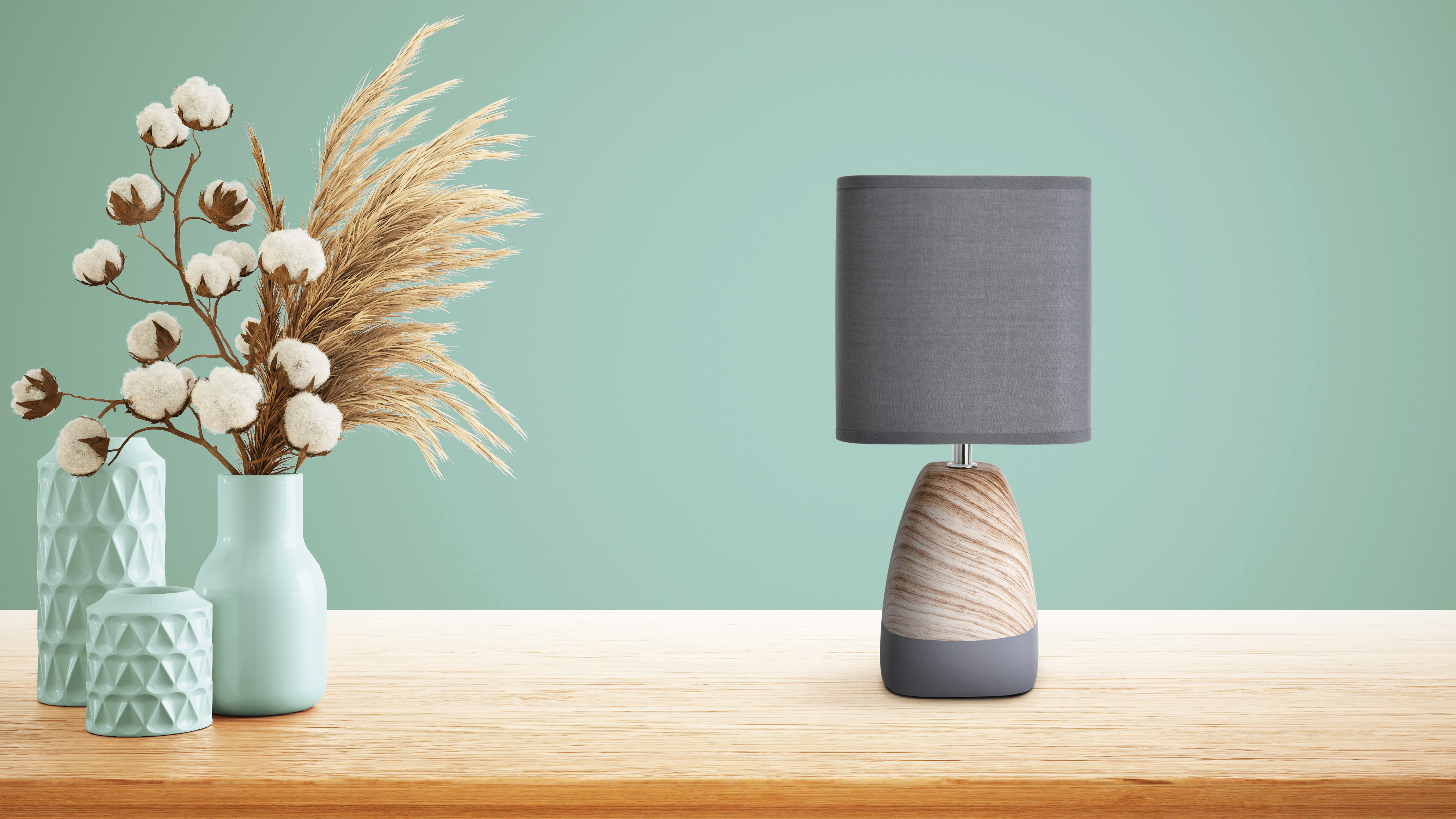 Eine Lampe mit grauem Schirm und Holzfuß neben einer Vase mit Baumwolle und Pampasgras.