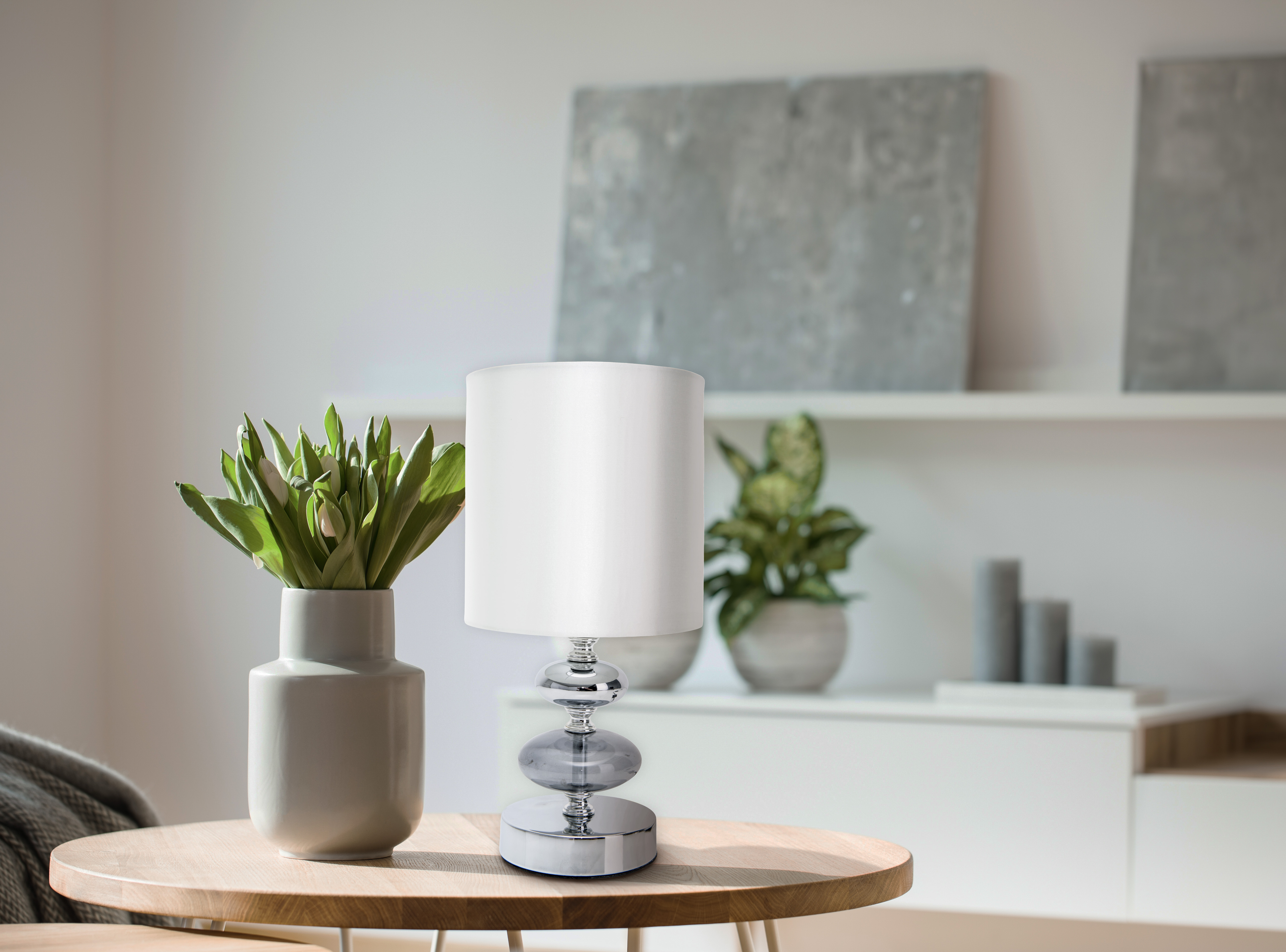Tischlampe mit weißem Schirm, Vase mit grünen Blättern, Hintergrund mit Dekoration.