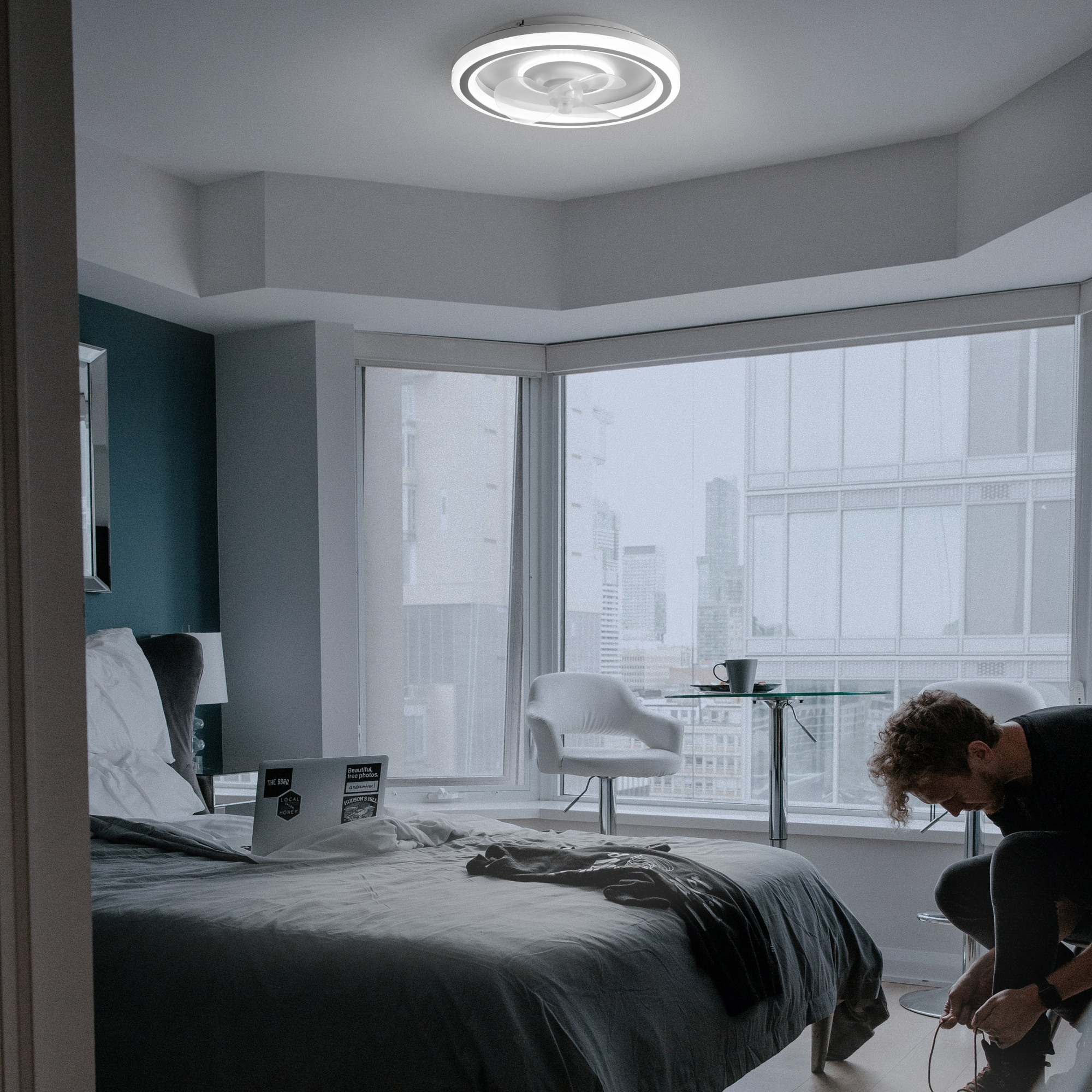Dormitorio con cama, vista de la ciudad. Una persona se ata los zapatos. Lámpara de techo, decoración blanca y azul.