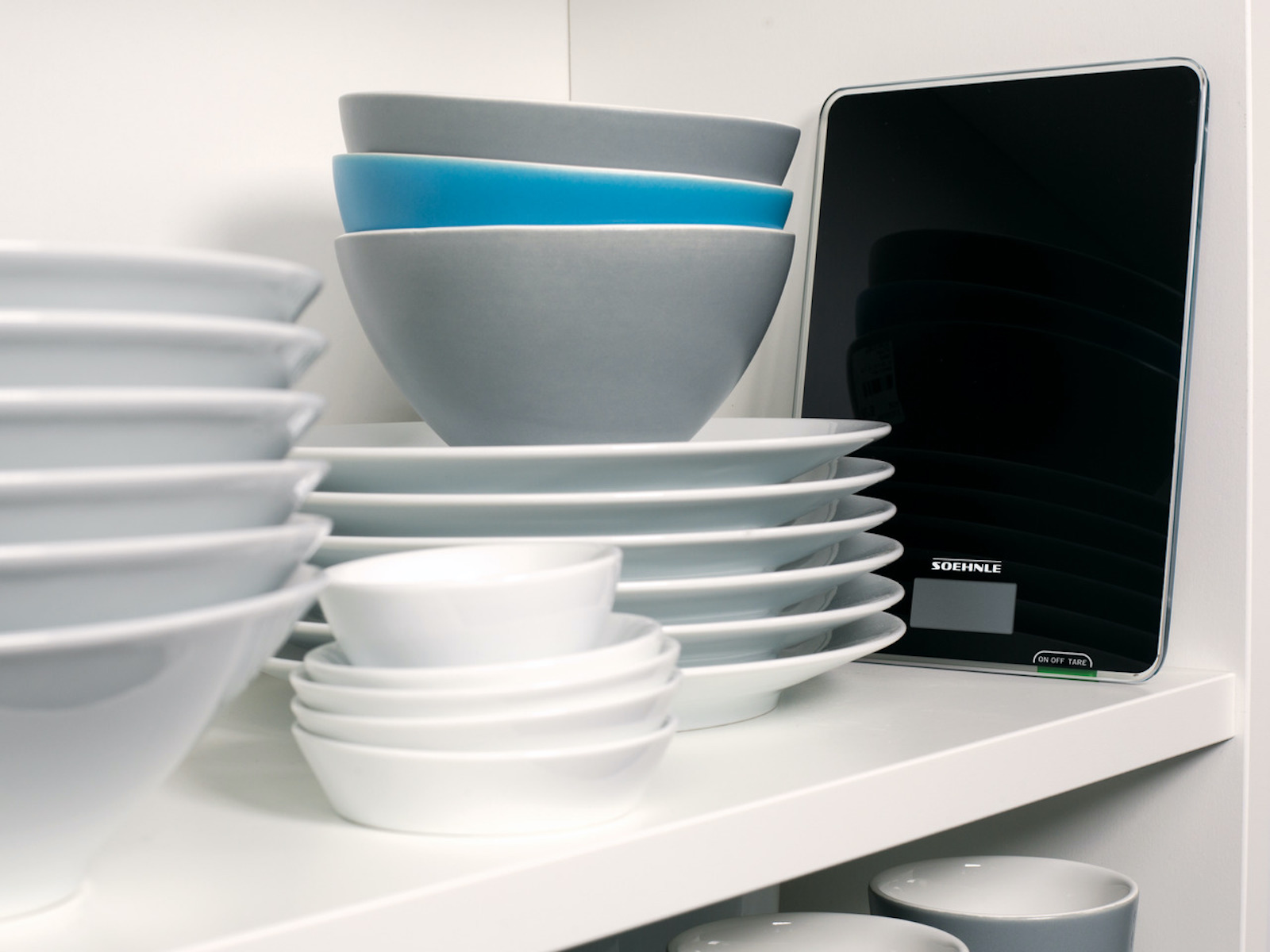 Czarna waga cyfrowa oraz stos misek i talerzy na białej półce. Białe tło.