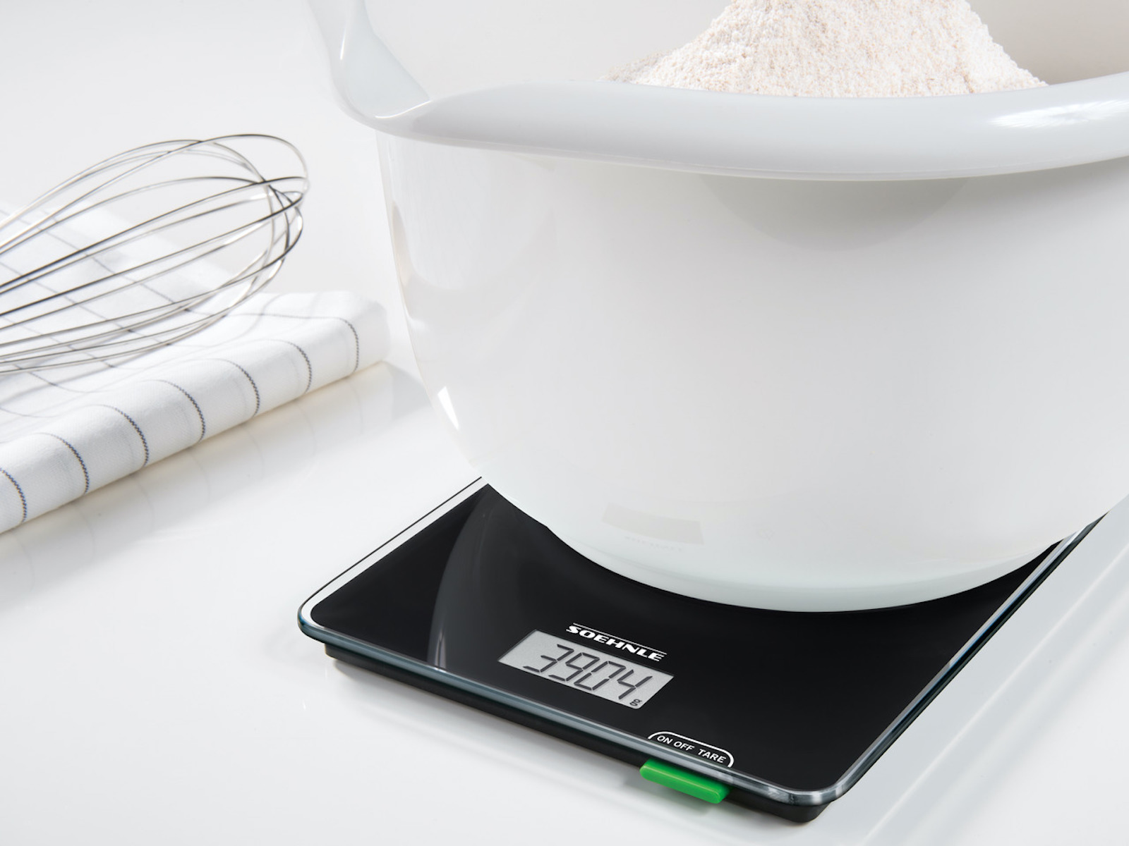 Biała miska na wadze wypełniona mąką, trzepaczka i ściereczka na białej powierzchni.