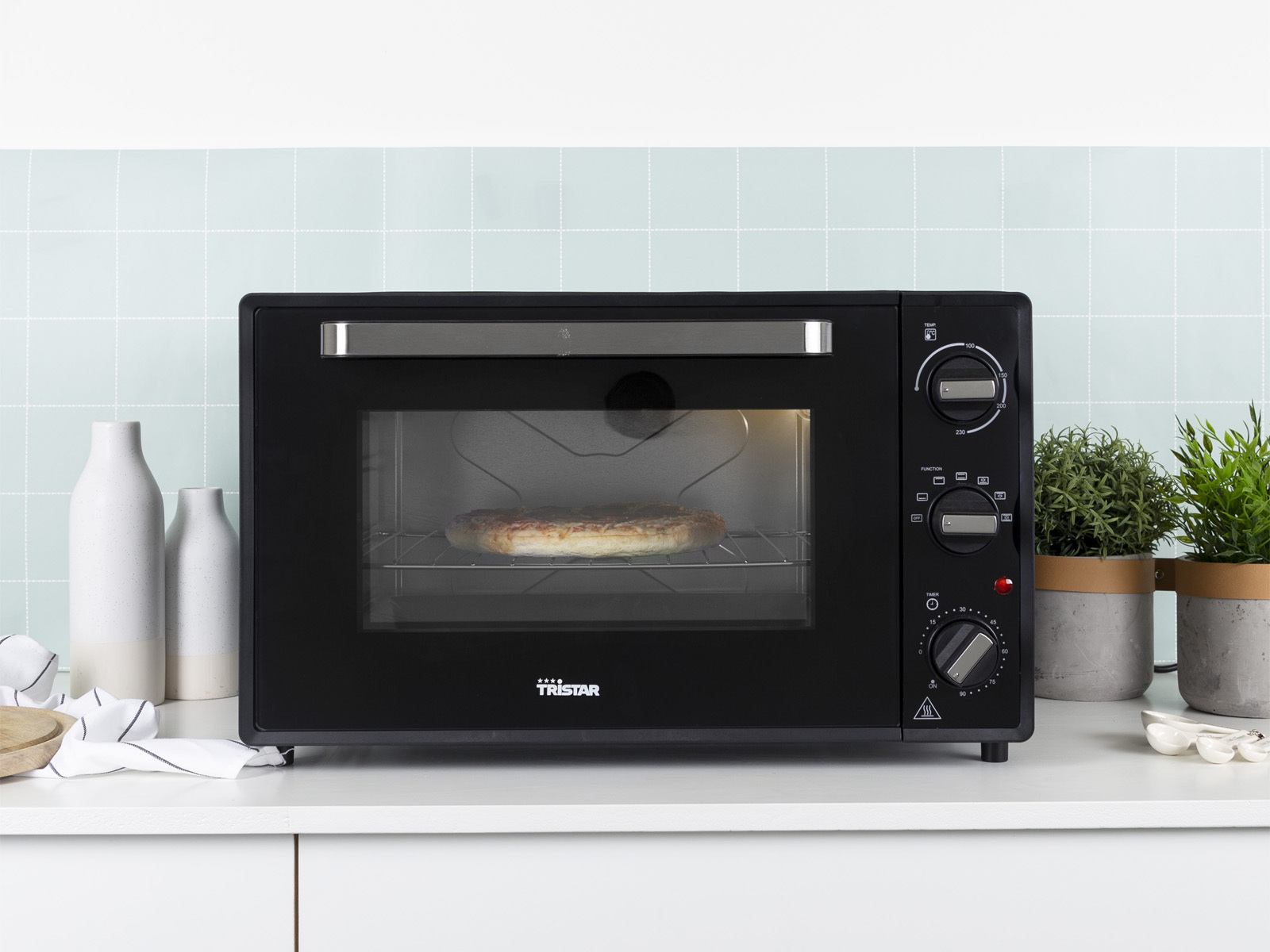 Un four noir avec une pizza sur un comptoir blanc. Des plantes et des vases sont à proximité.