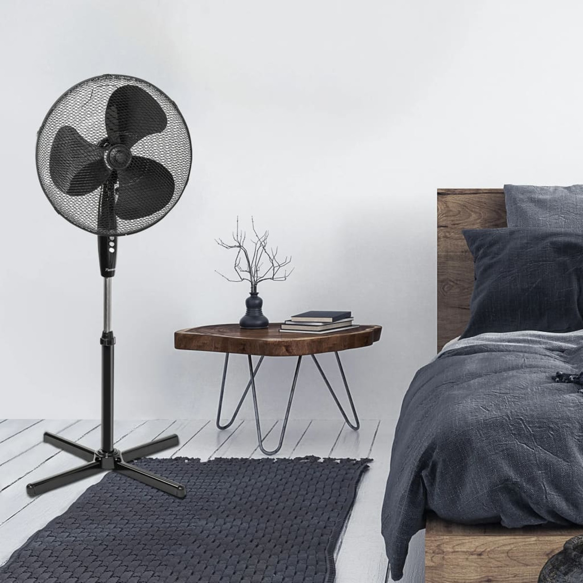Czarny wentylator stojący z siatkową klatką. Stoi na czarnej podstawie. Drewniany stolik i łóżko w tle.