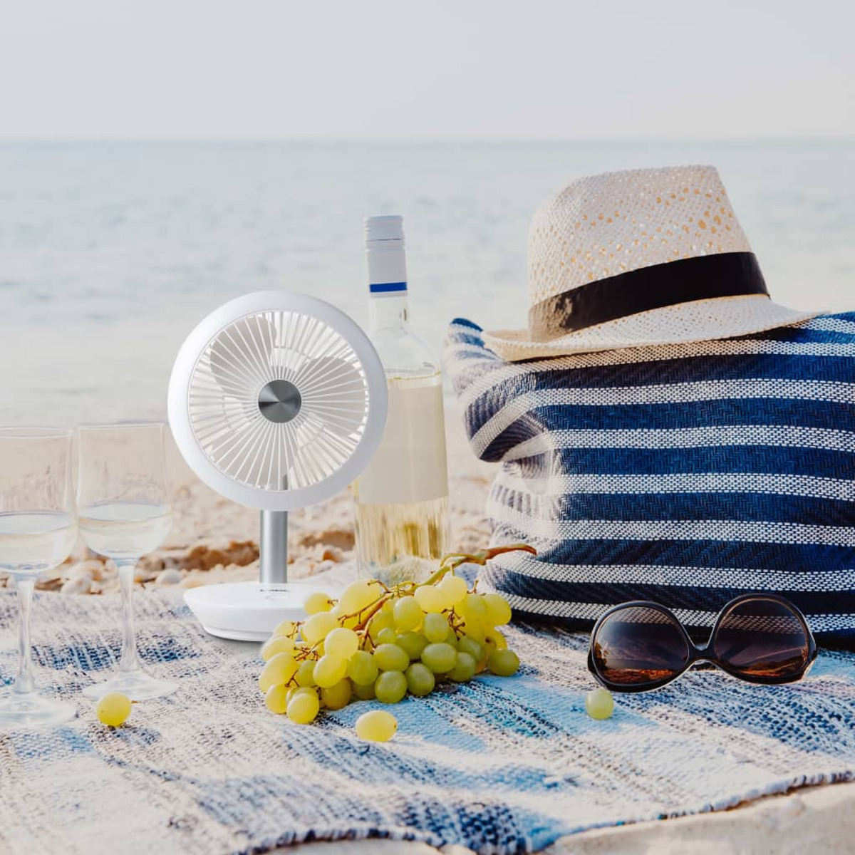 Strandszene mit Ventilator, Wein, Trauben, Hut und Sonnenbrille. Blaue Streifentasche auf Sand.