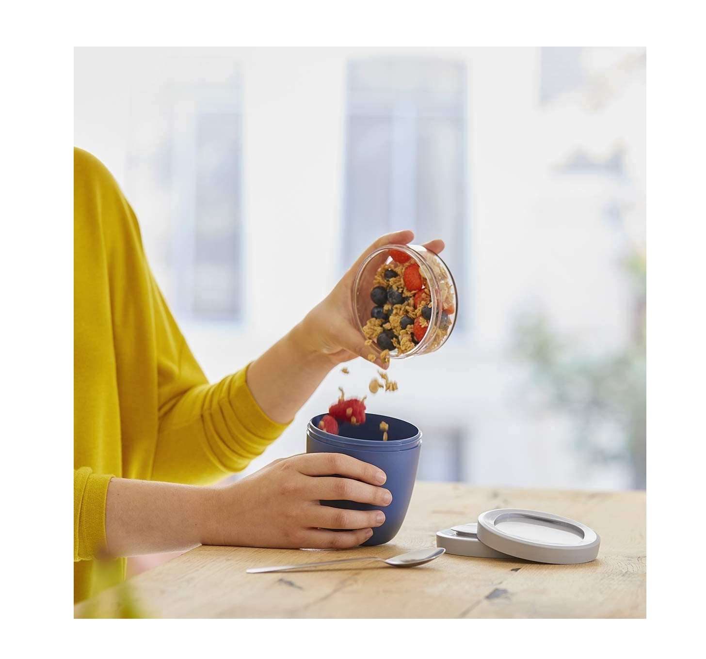 Eine Person gießt Müsli und Beeren aus einem Glasbehälter in einen blauen Behälter auf einem Holztisch.