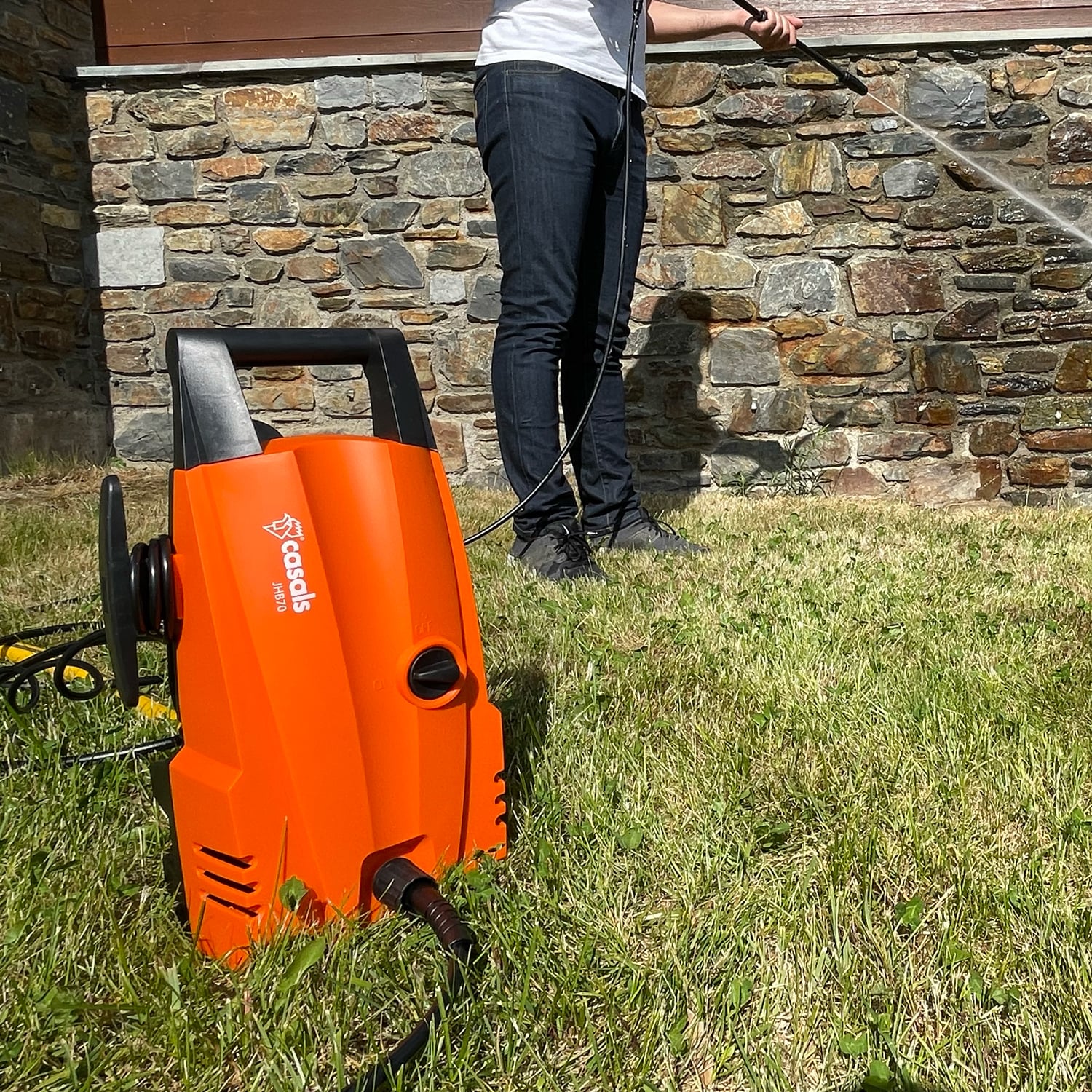 Una hidrolimpiadora naranja está sobre la hierba. Persona lava pared de piedra con manguera de agua.