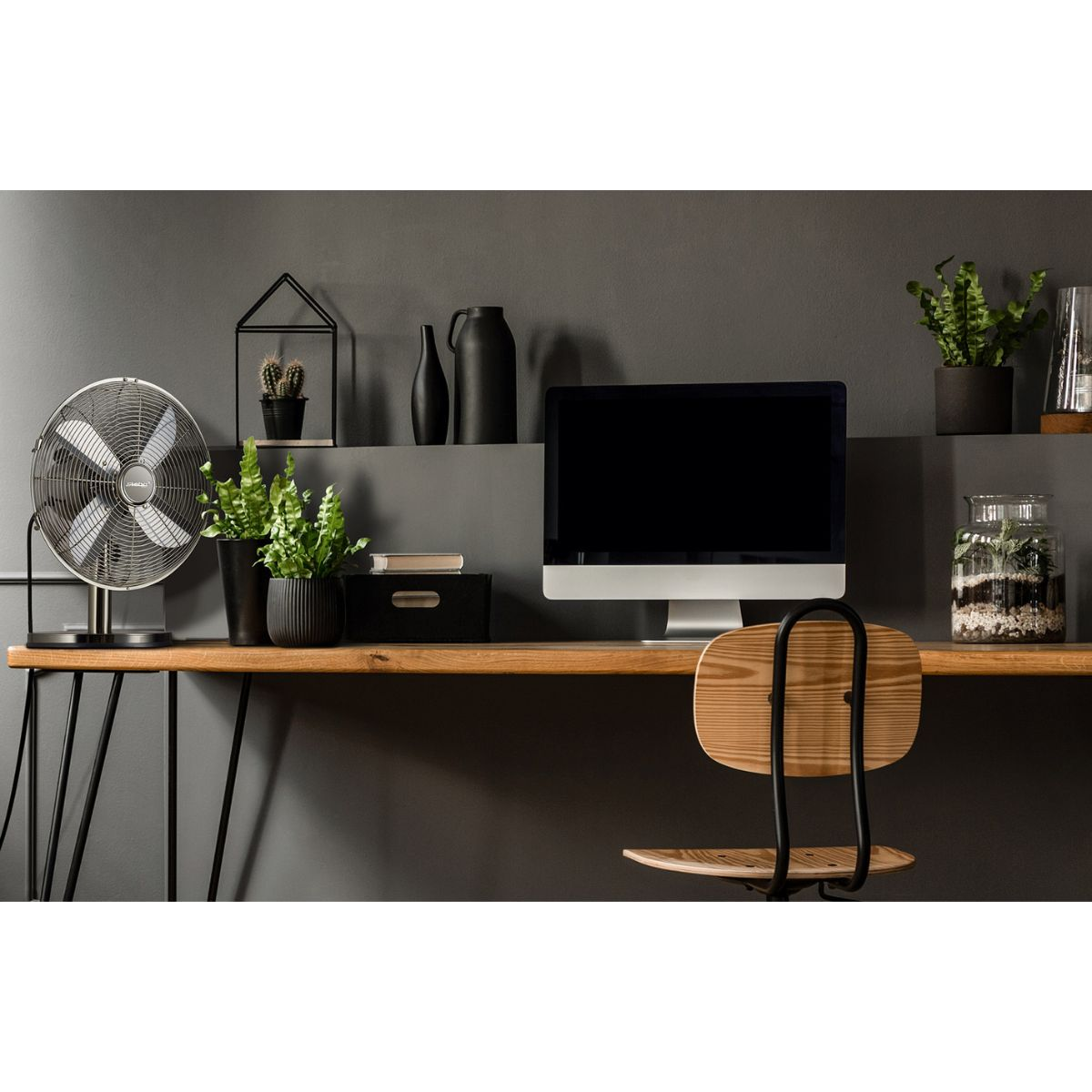 Escritorio con ventilador, monitor, plantas y silla. Pared gris oscuro en el fondo.
