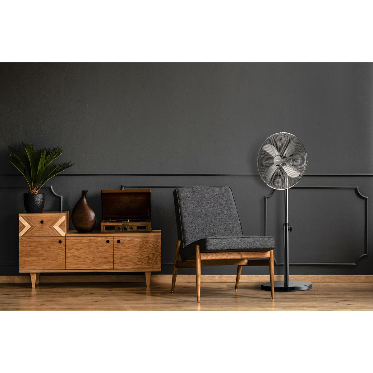 Una habitación con un mueble de madera, sillón y ventilador de pie. La pared es gris oscuro.