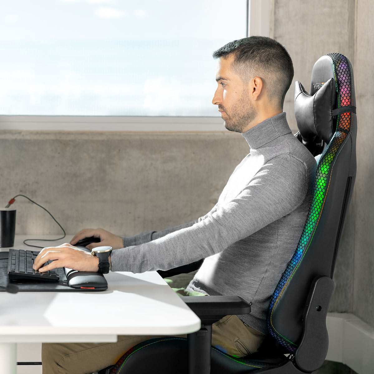 Hombre en silla gaming, usando teclado y ratón. Escritorio blanco. Fondo ventana y pared.