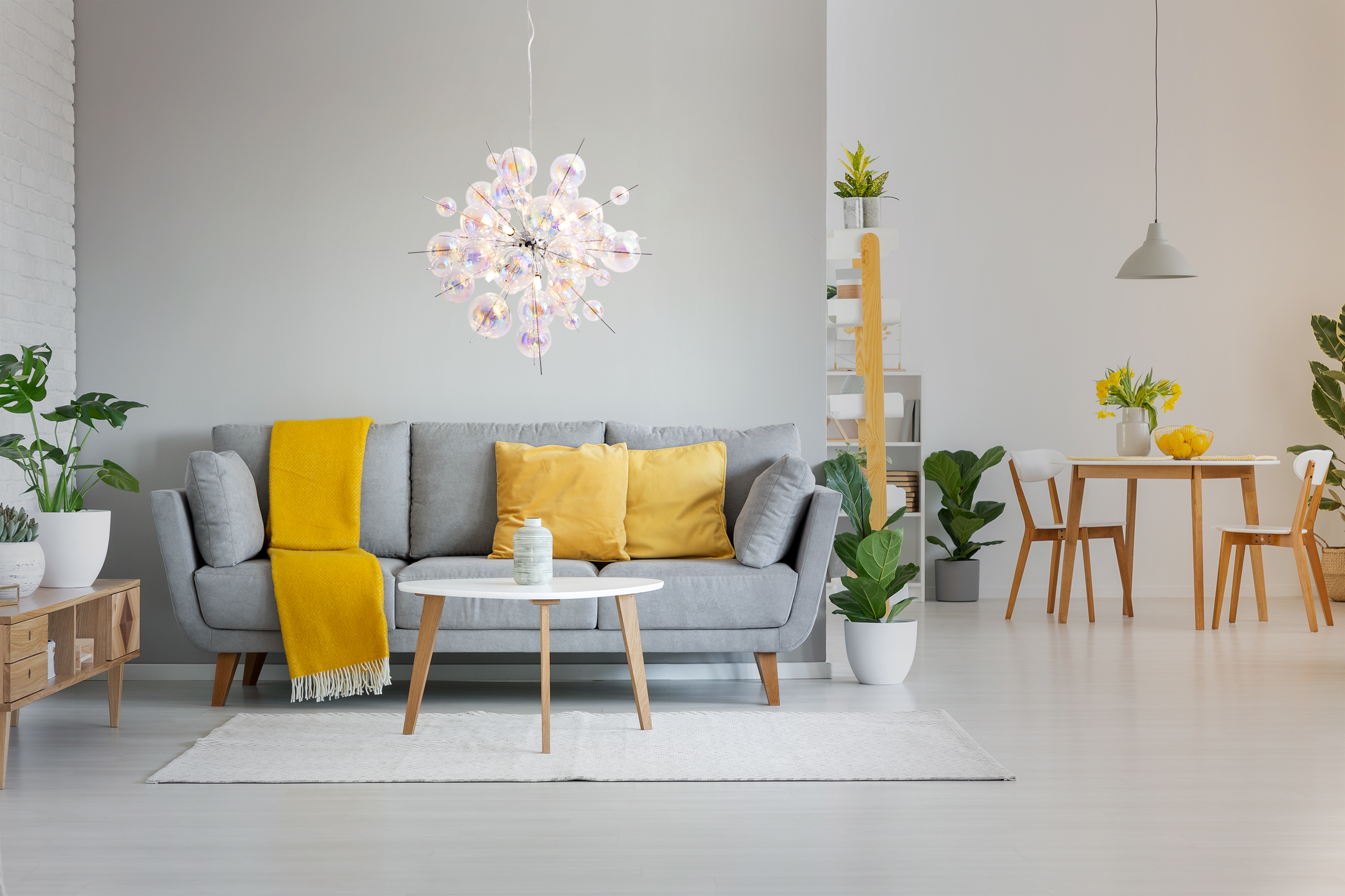 Modernes Wohnzimmer mit grauem Sofa, gelben Akzenten und weißem Couchtisch. Ein Kronleuchter hängt darüber.