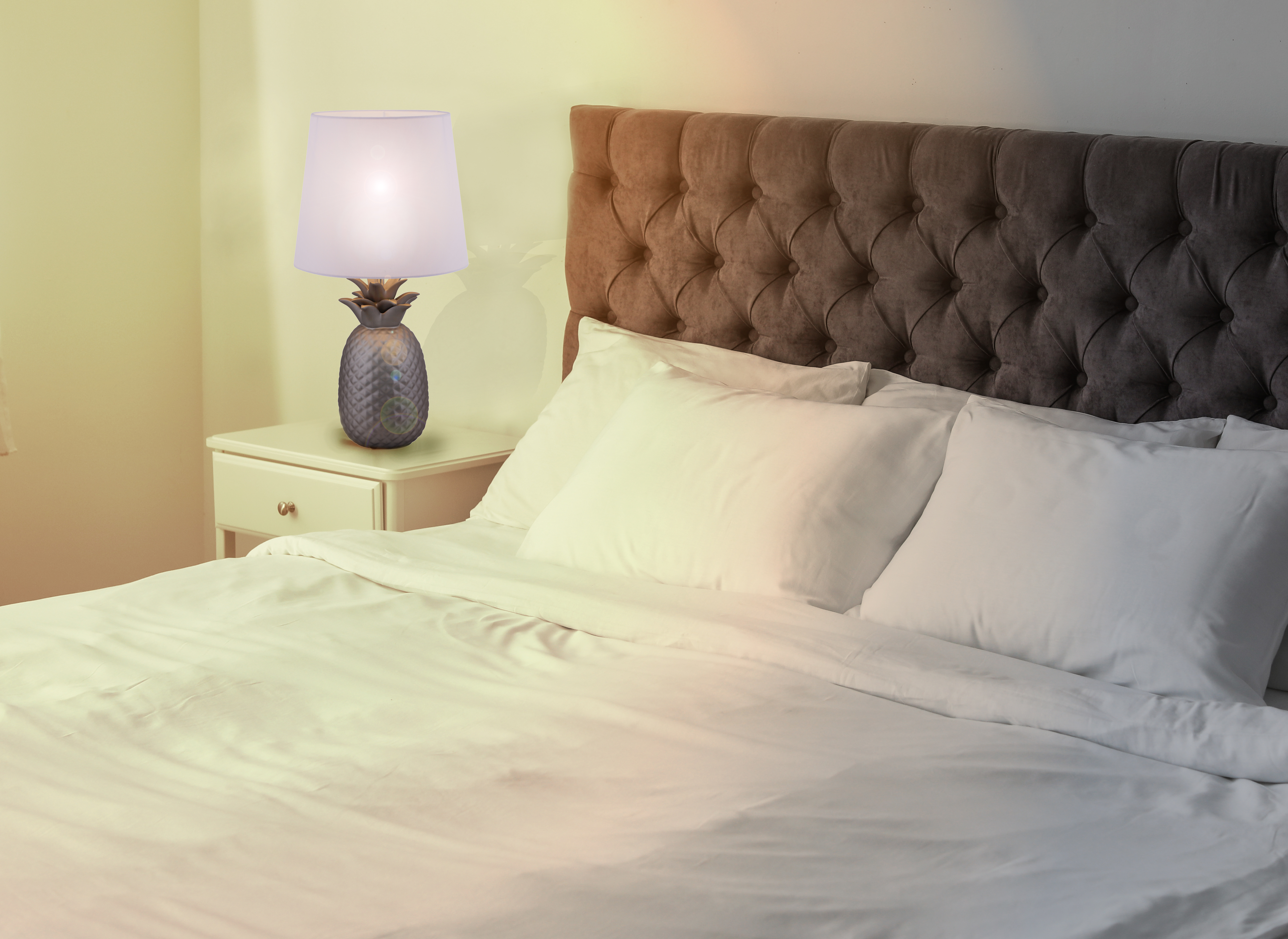 Schlafzimmer mit Ananas-Lampe auf einem Nachttisch. Bett mit weißen Laken und Kissen.