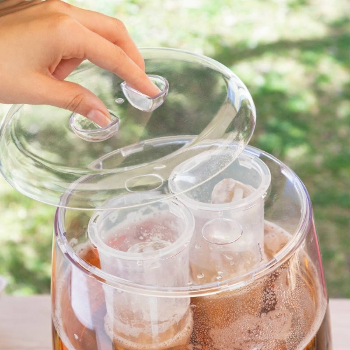 Una mano che apre un contenitore di plastica trasparente pieno di ghiaccio. Il contenitore si trova in un bicchiere pieno di birra.
