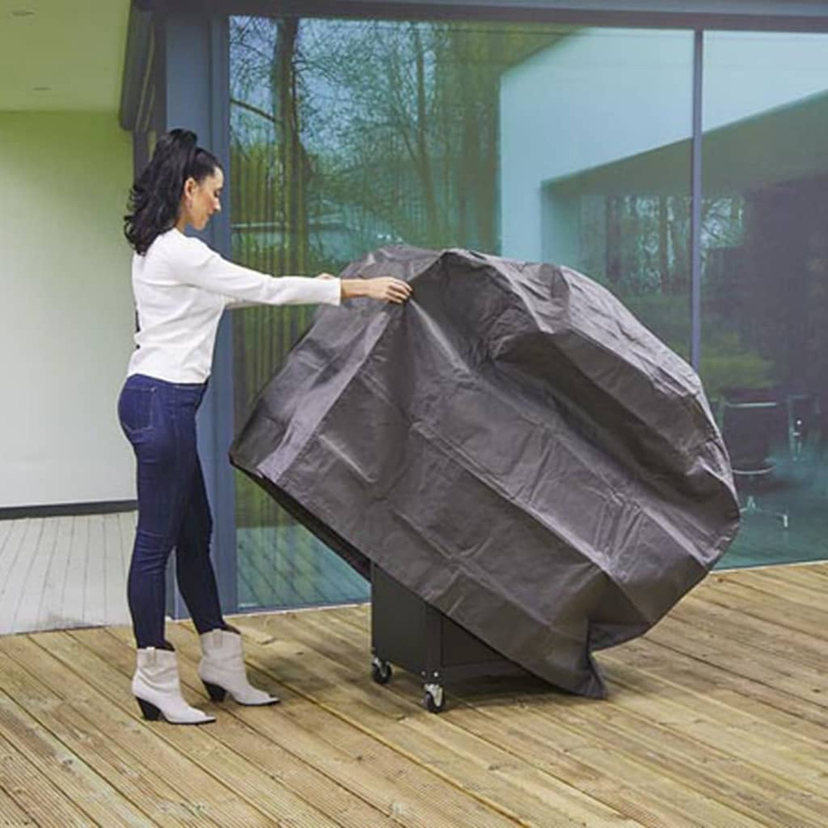 Frau, die einen Grill mit einer schwarzen Abdeckung auf einer Holzterrasse abdeckt, im Freien.