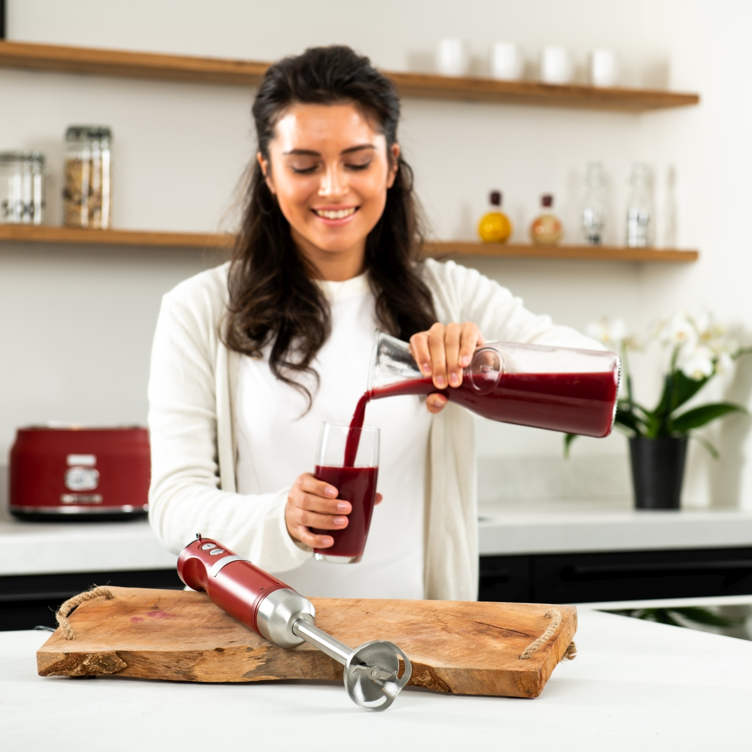 Frau gießt roten Saft in ein Glas mit einem roten Handmixer auf einem Holzbrett.