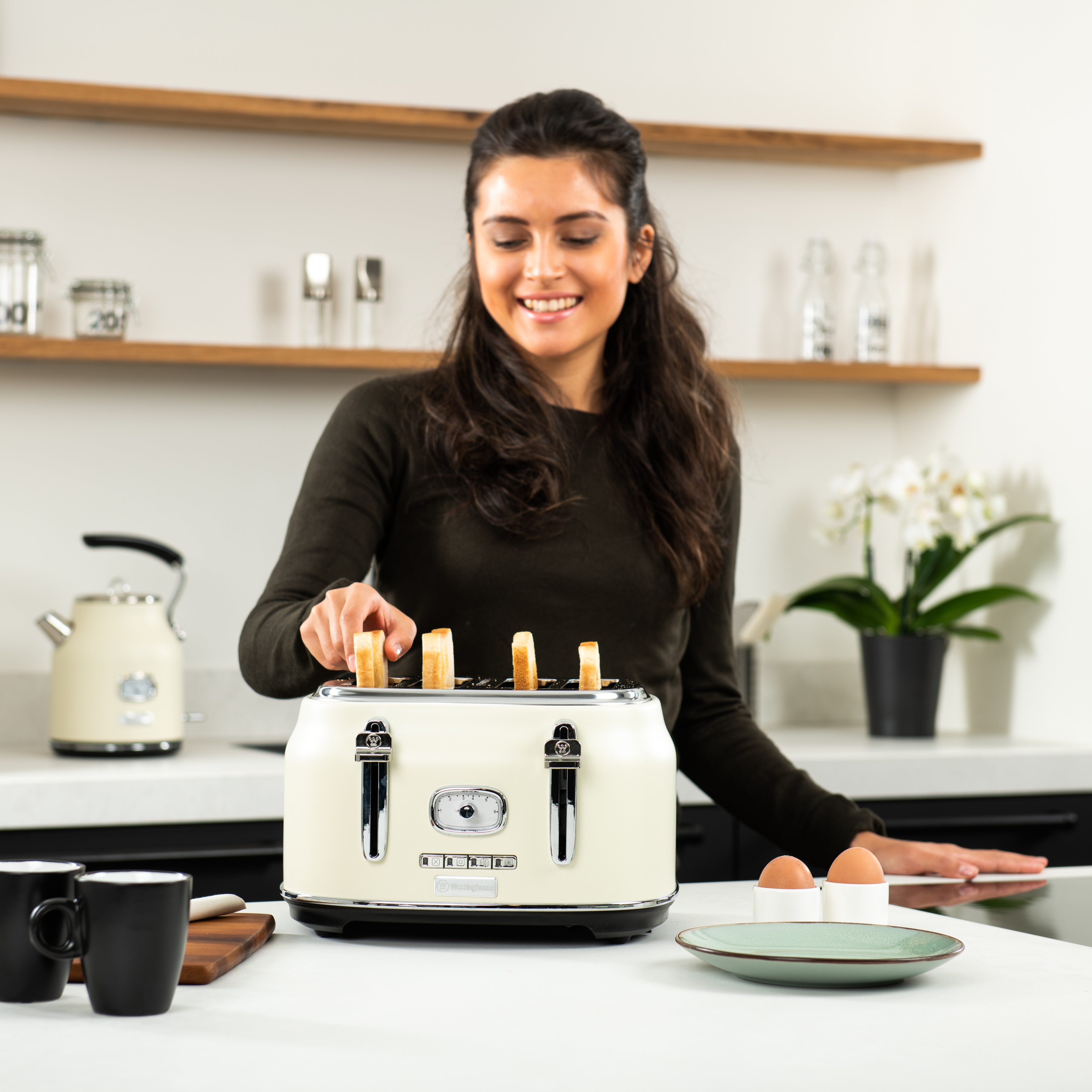 Eine Frau legt Toast in einen cremefarbenen Toaster. Es gibt Eier, einen Wasserkocher und Blumen.