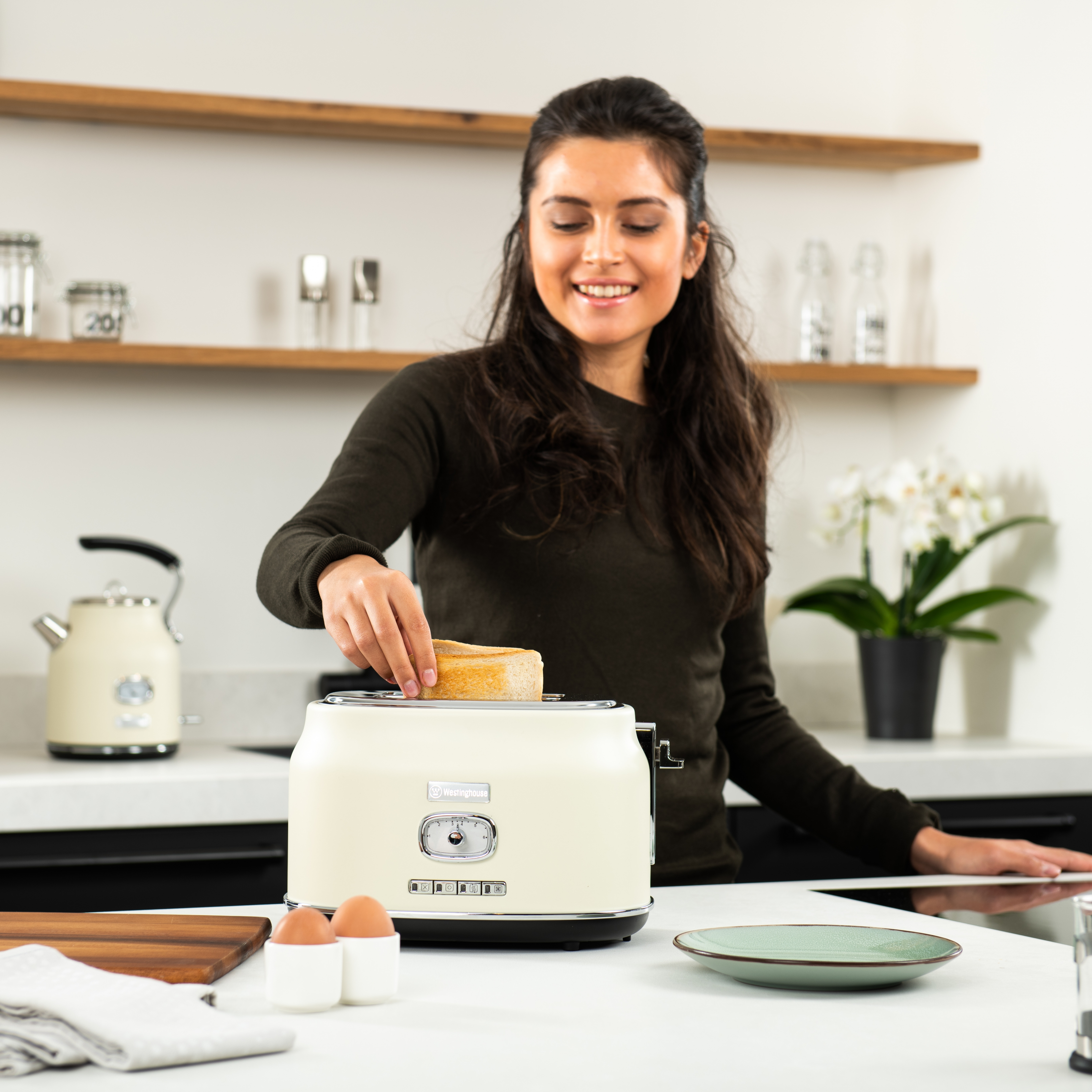 Frau legt Toast in einen cremefarbenen Toaster. Küchenhintergrund mit Wasserkocher, Eiern und einem Teller.