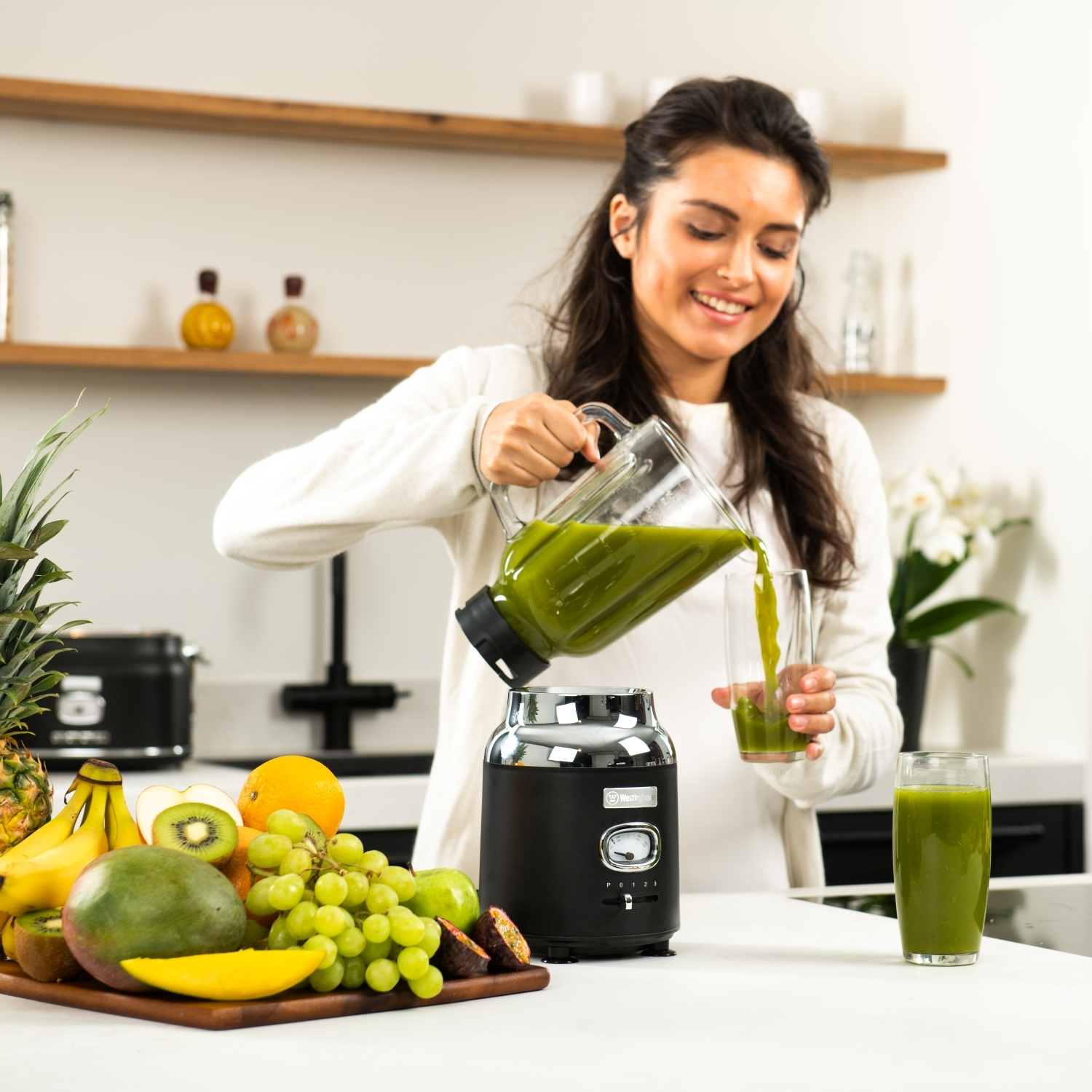 Frau gießt grünen Saft aus einem Mixer in ein Glas. Obst auf der Theke.