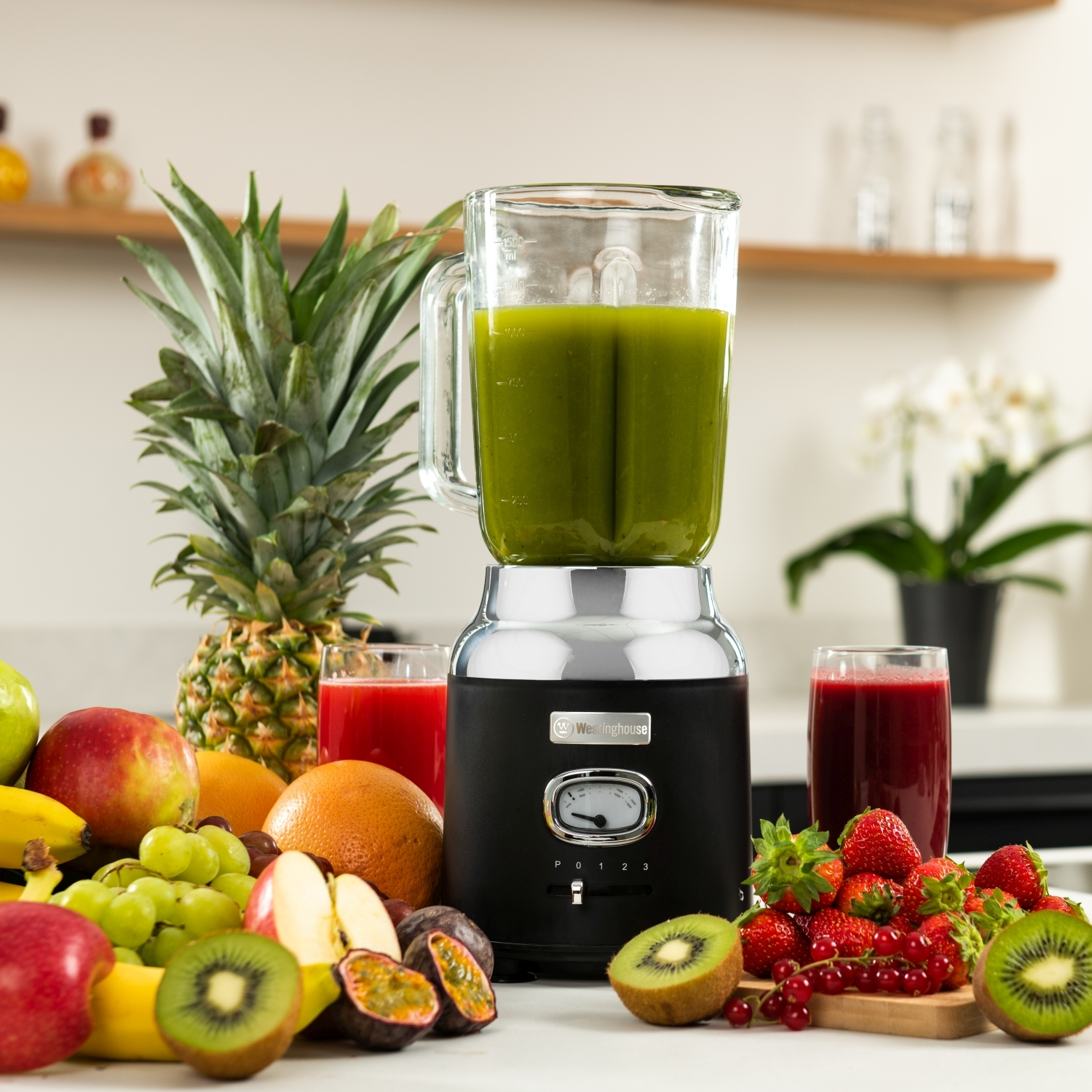 Mixer mit grünem Smoothie, Früchten und zwei Gläsern Saft. Ananas, Äpfel und Kiwis liegen auf dem Tisch.