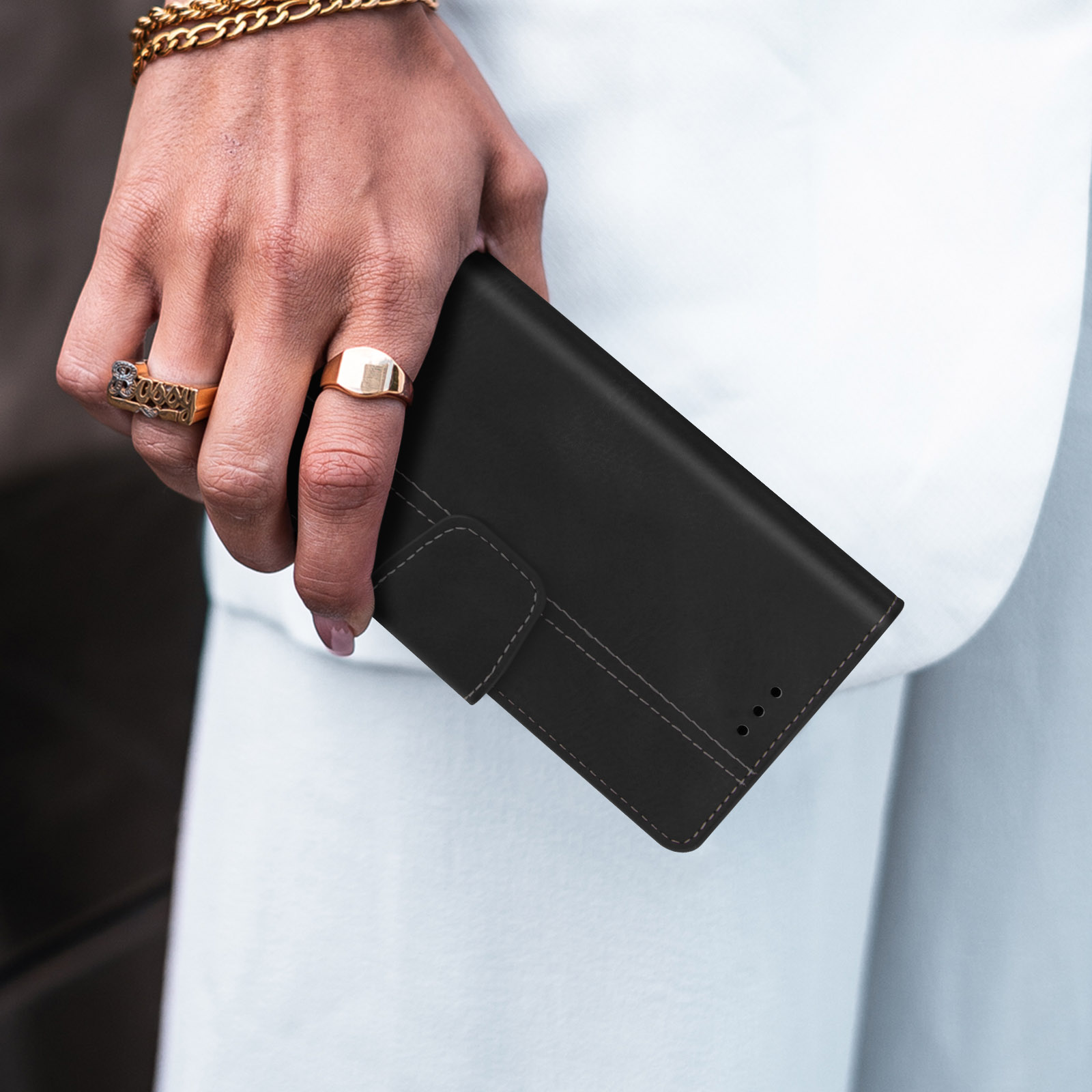 Eine Hand hält eine schwarze Brieftasche, mit goldenen Ringen an den Fingern, vor einem weißen Mantel.