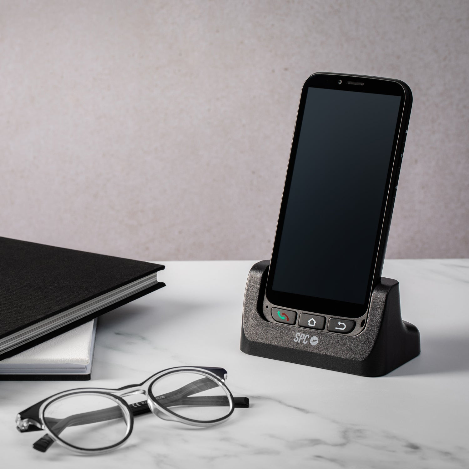 Un smartphone en una base de carga, gafas y un libro sobre una superficie de mármol.