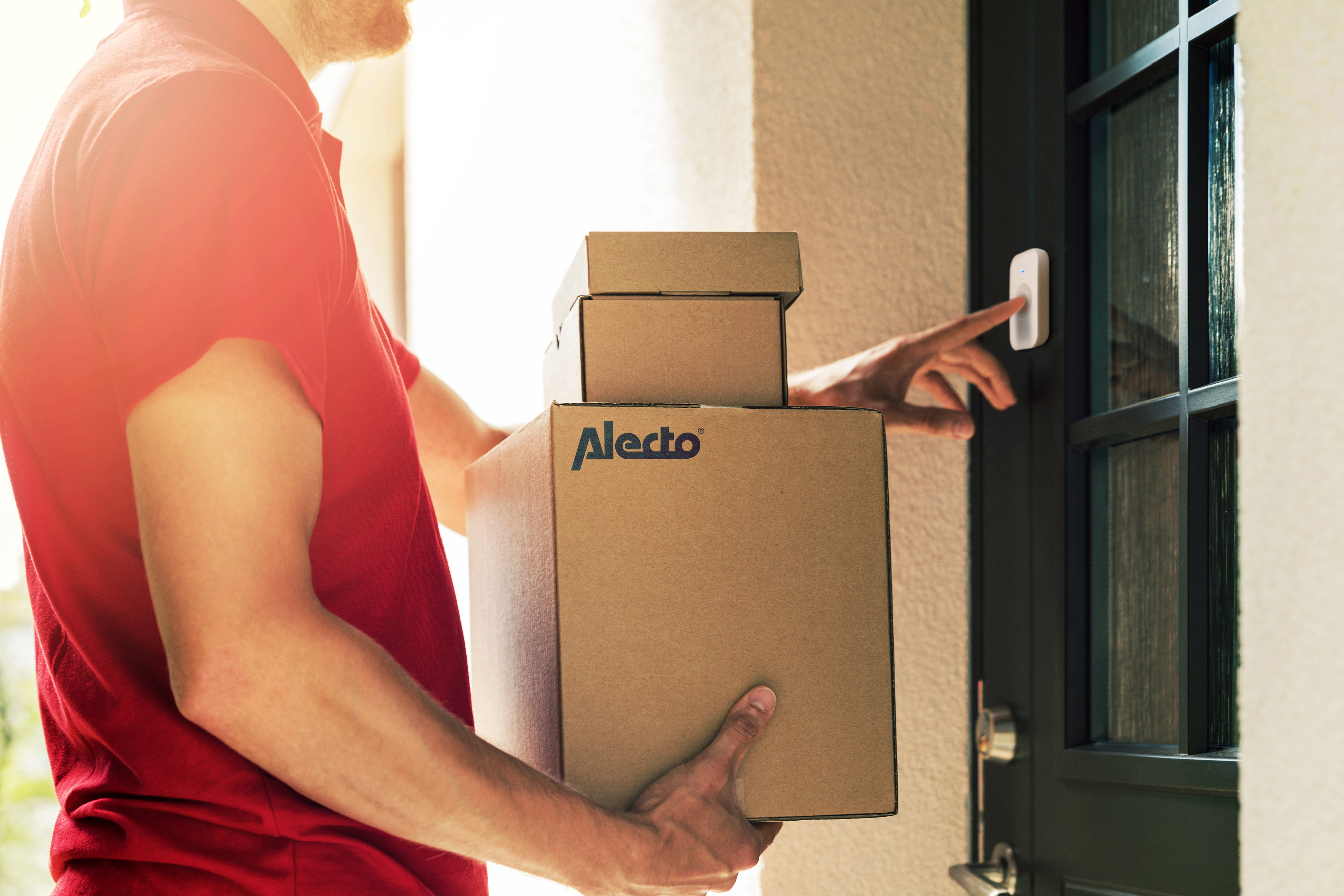 Un livreur en chemise rouge sonne à la porte tout en tenant des boîtes avec le logo Alecto.