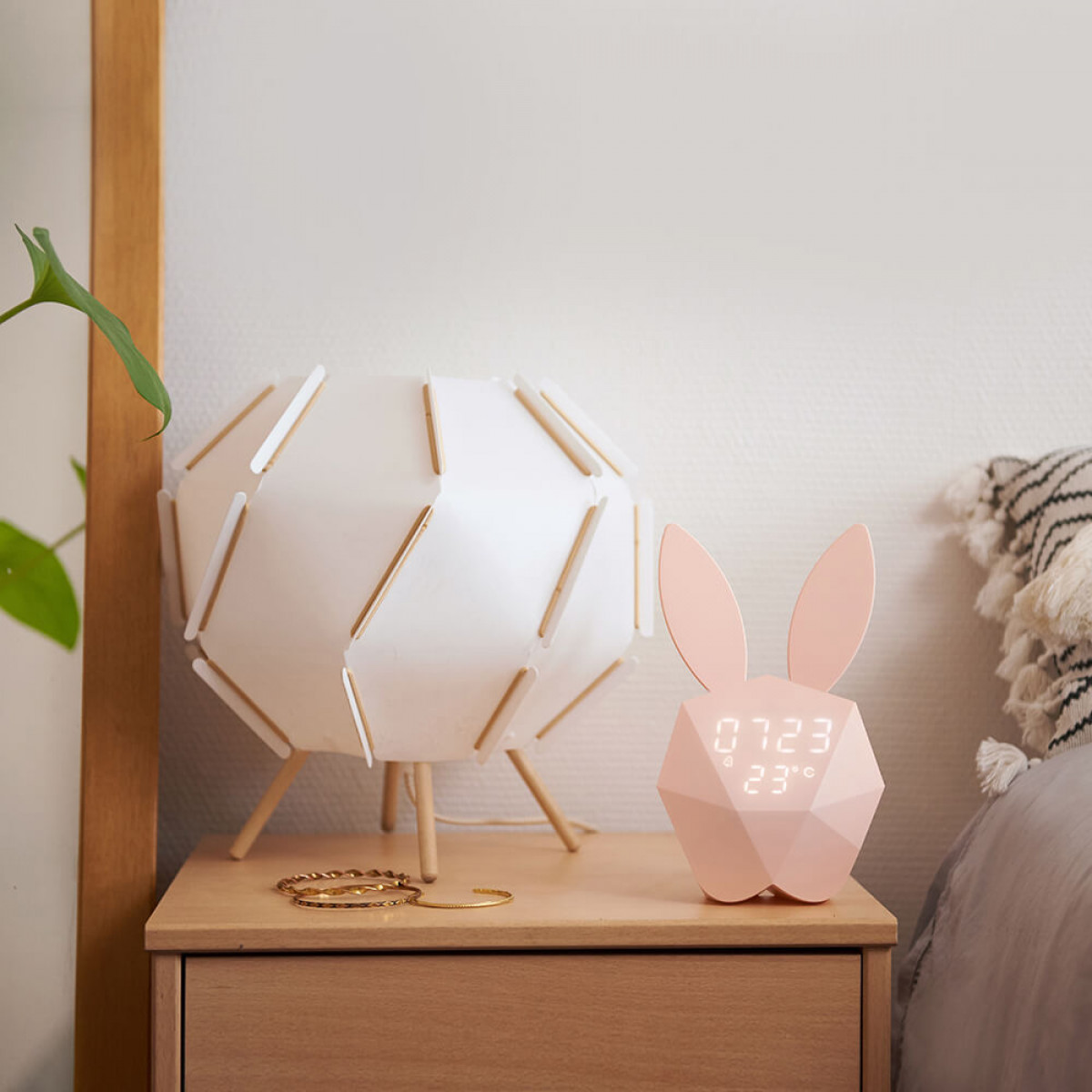 Ein Nachttisch mit Lampe, einem hasenförmigen Wecker und einem Bett im Hintergrund.