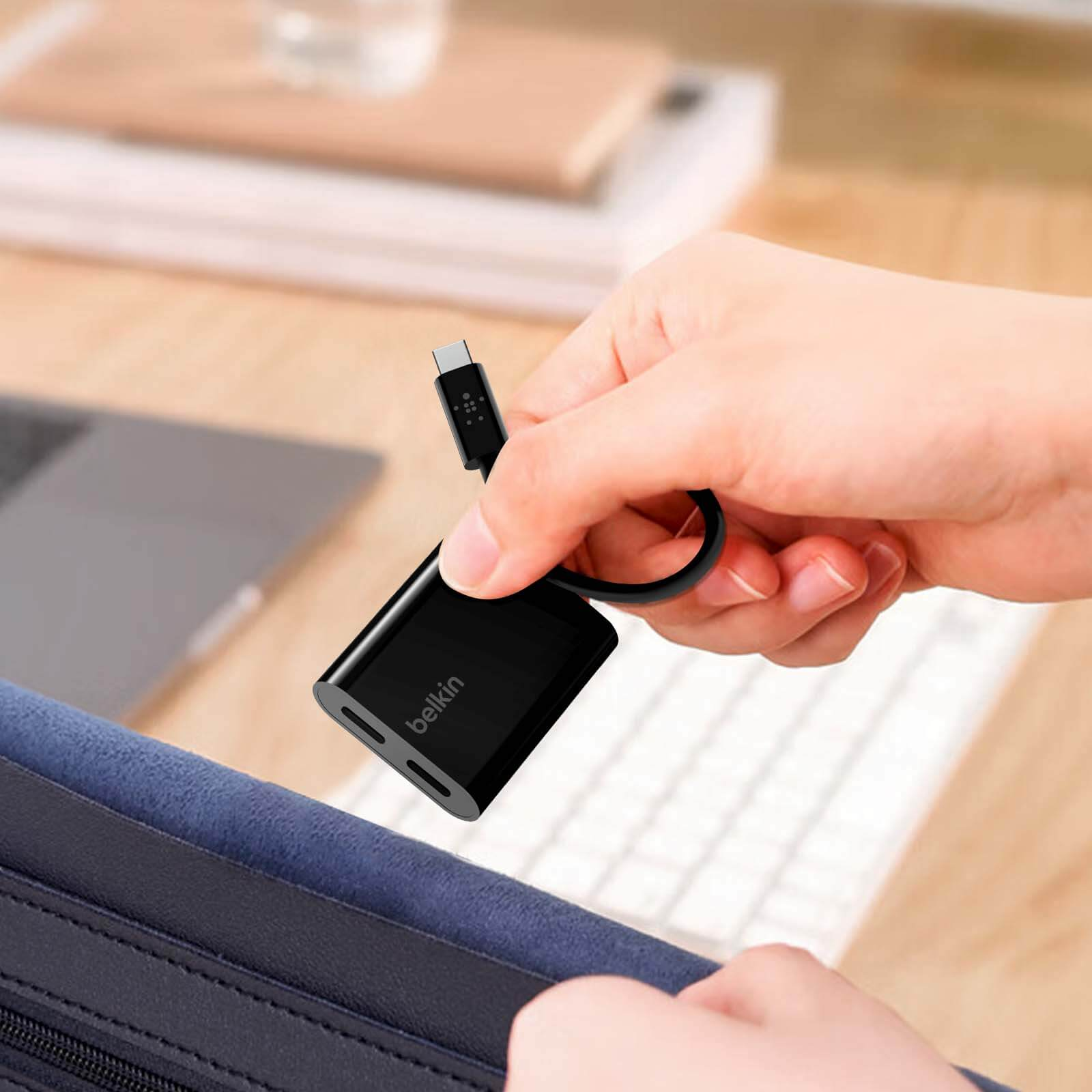 Mano che tiene un adattatore Belkin nero collegato a un laptop, su uno sfondo chiaro.