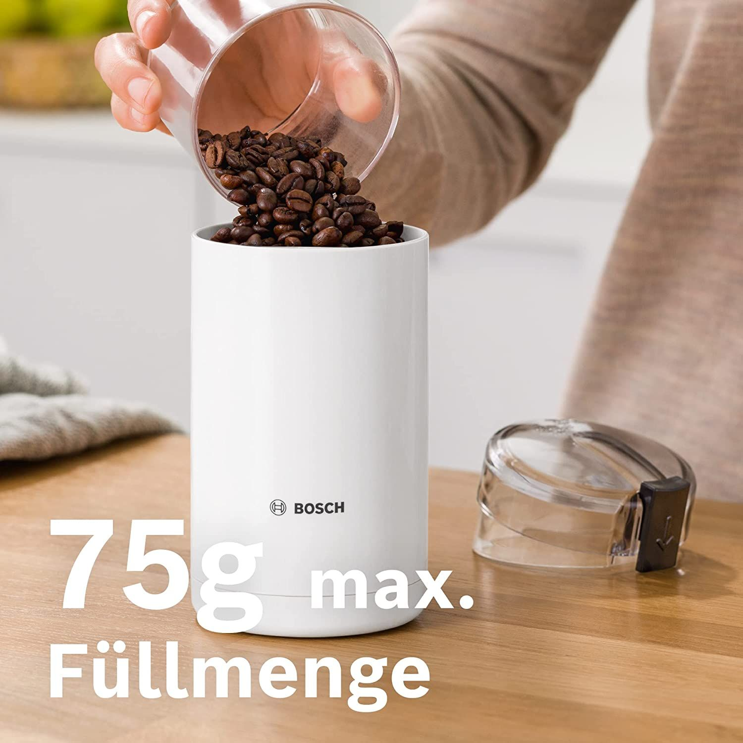 Eine weiße Kaffeemühle wird mit Kaffeebohnen gefüllt. Ein transparenter Deckel ist in der Nähe.