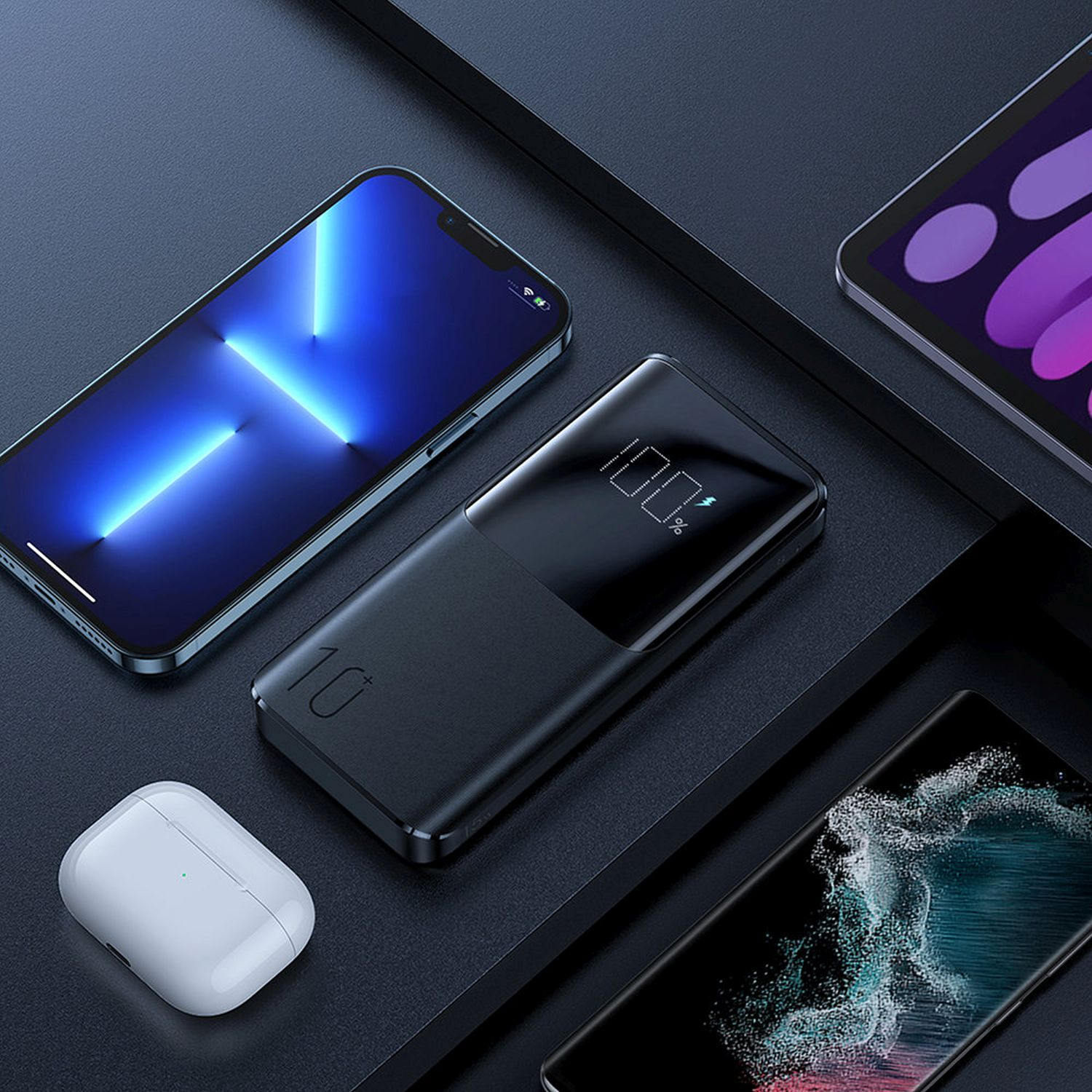 Schwarze Powerbank mit Digitalanzeige neben Telefon, Tablet und Ohrhörern auf dunkler Oberfläche.