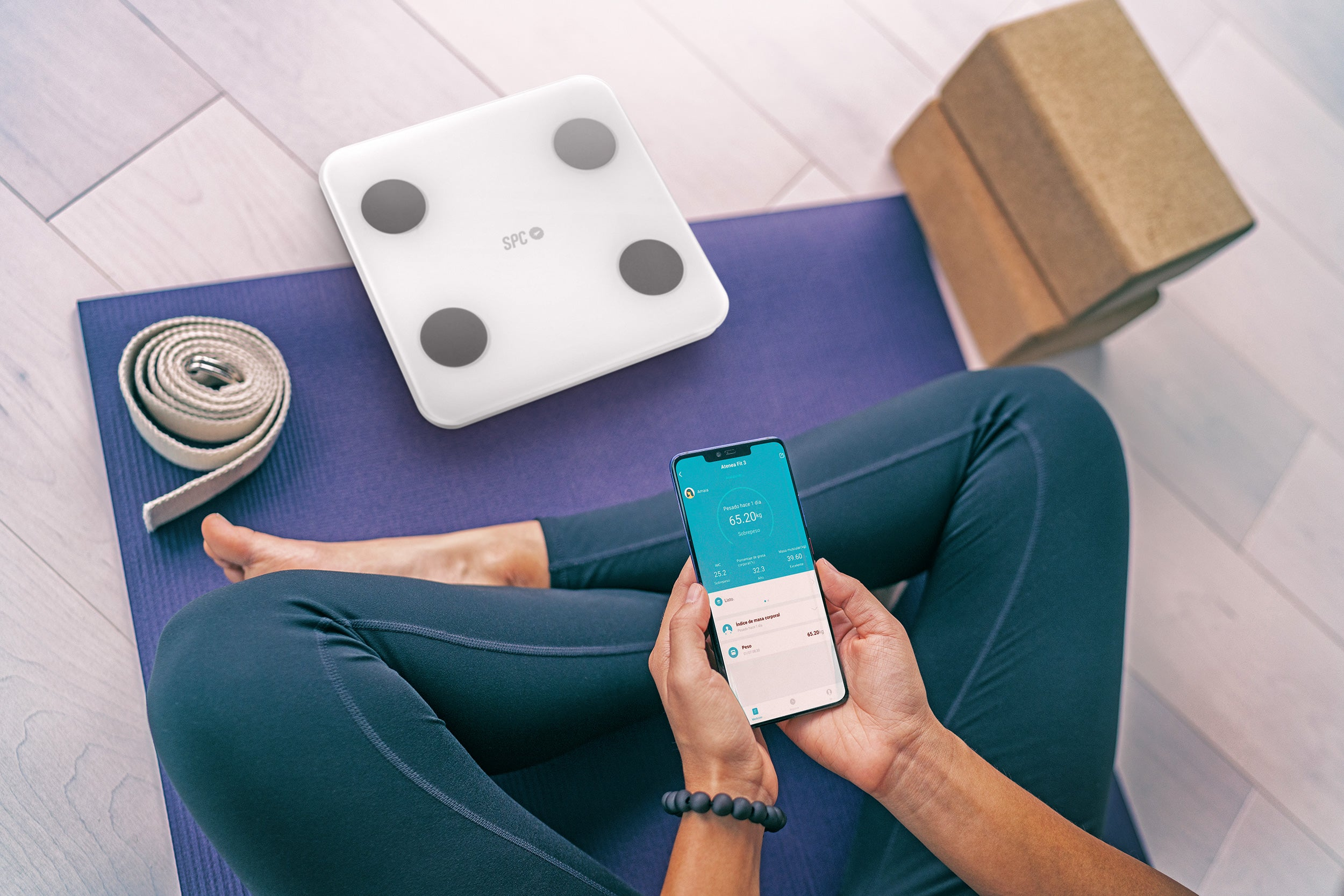 Osoba używa wagi i telefonu do pomiaru masy ciała, z matą do jogi w tle.