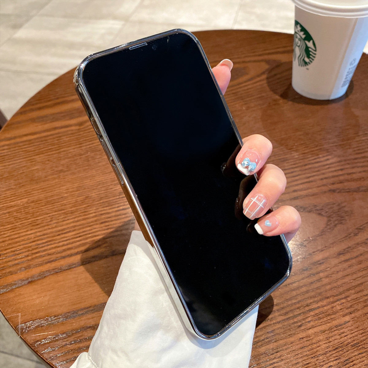 Hand hält ein schwarzes Smartphone in einem silbernen Gehäuse. Im Hintergrund ein Kaffeebecher und Wand.