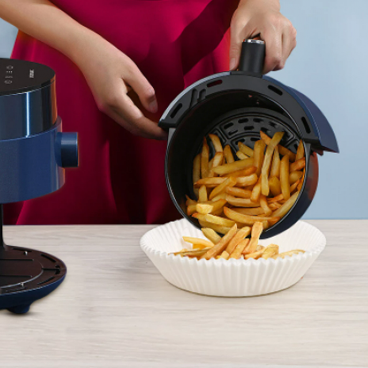 Person pouring fries from a blue air fryer into a paper bowl. Light wooden surface.