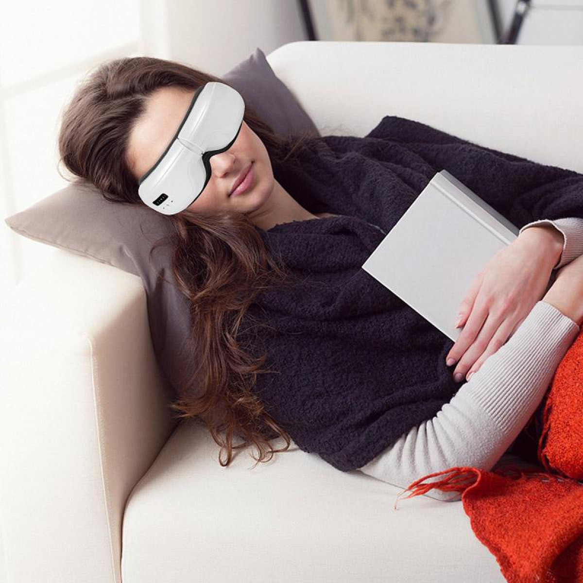 Frau mit Augenmaske auf Couch, hält Buch. Eine orangefarbene Decke ist auch auf der Couch.