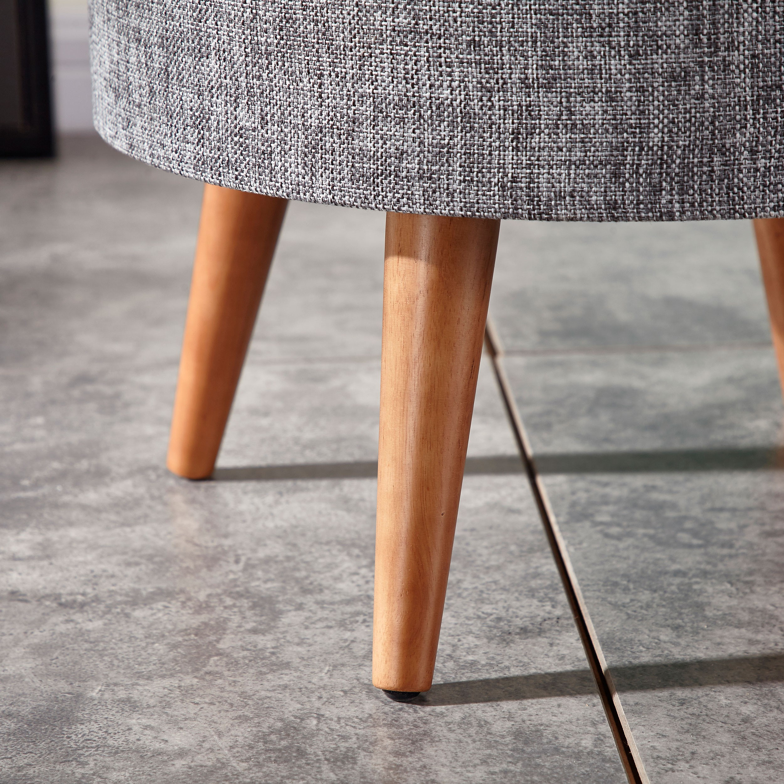 Primer plano de taburete. Parte superior de tela gris con patas de madera en suelo embaldosado.