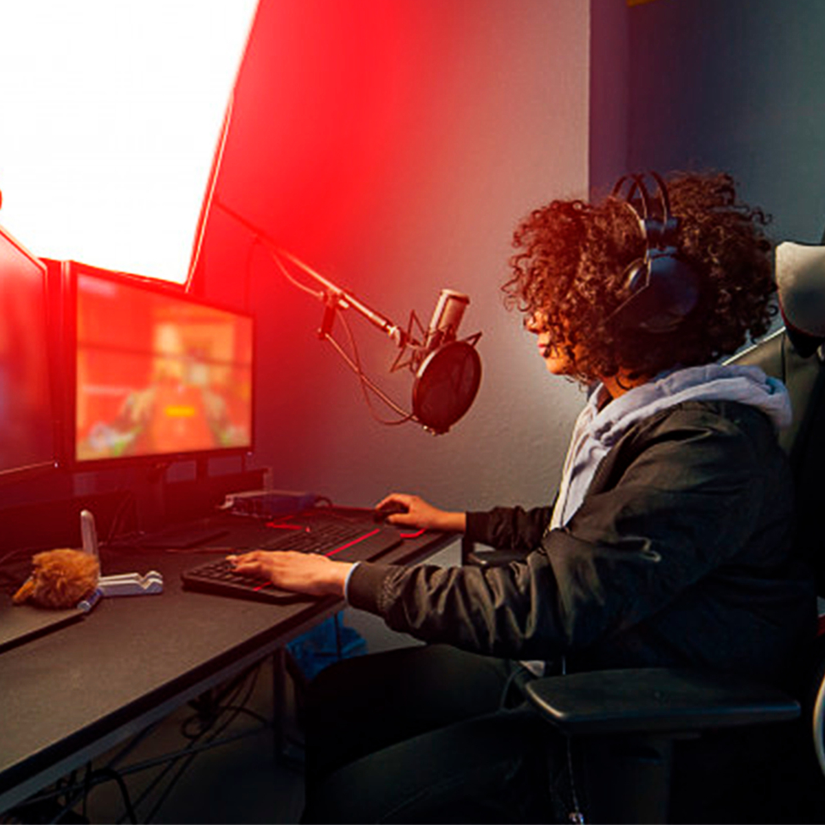 Mujer en configuración de juegos, usando teclado y ratón. Monitores y luz roja en el fondo.
