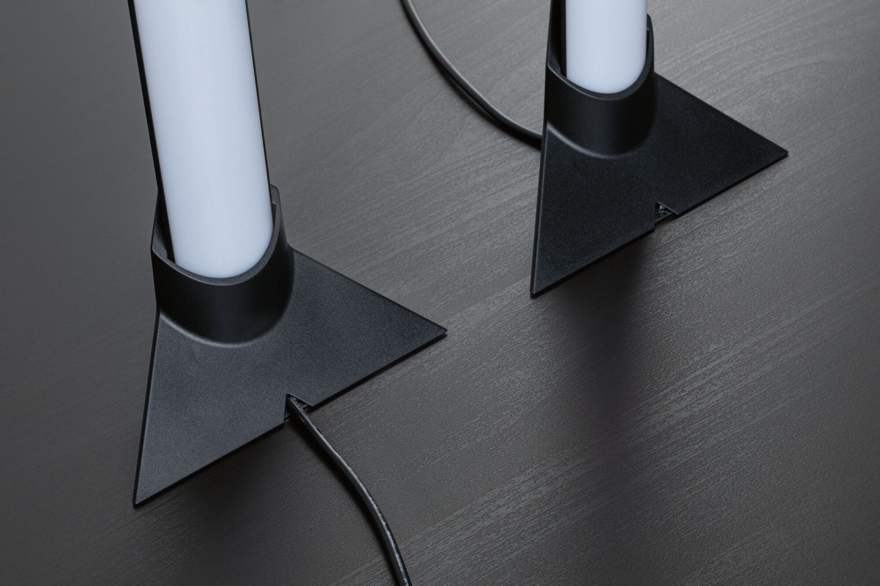 Due lampade moderne con tubi bianchi e basi triangolari nere su superficie in legno scuro.