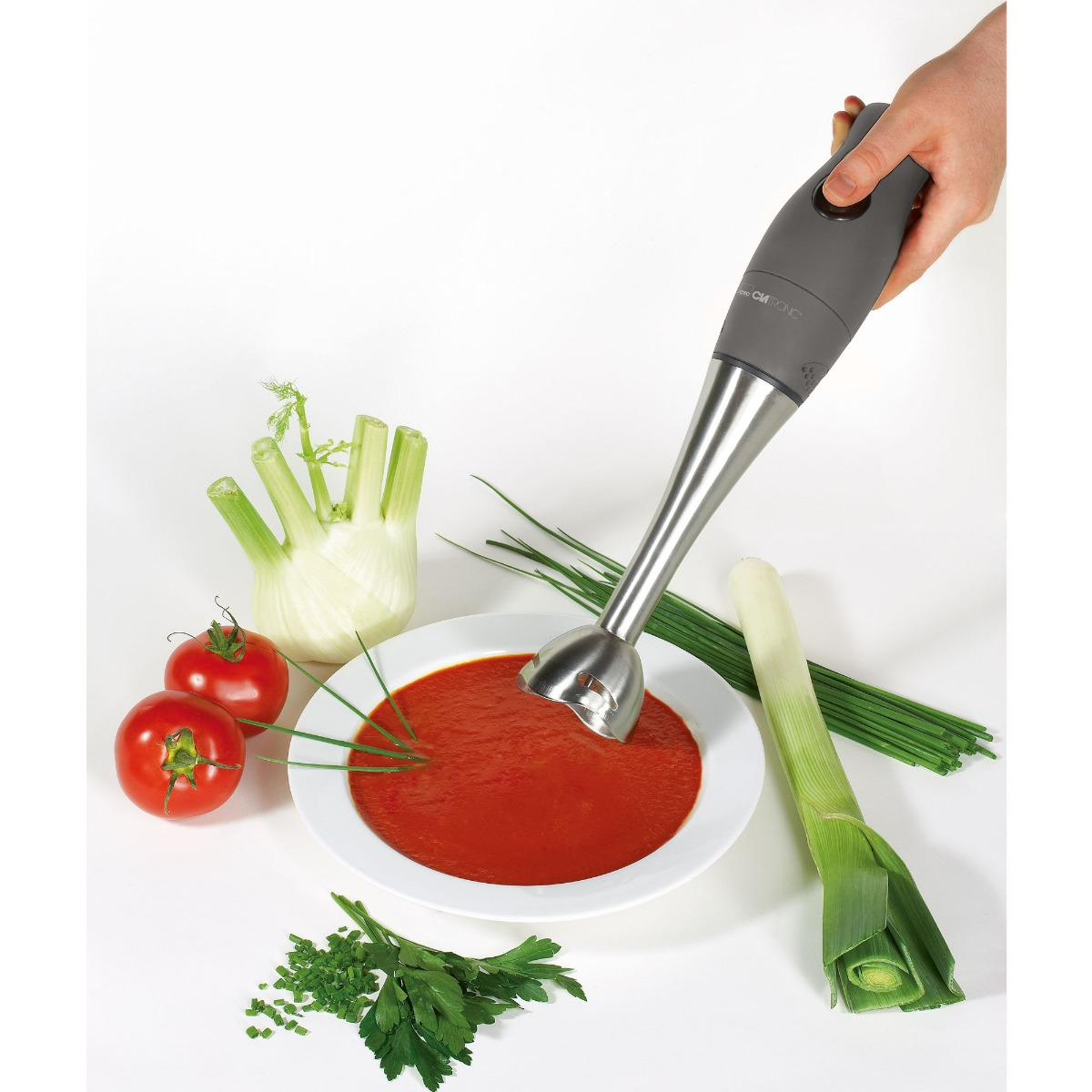 Dłoń trzymająca blender ręczny miksujący zupę w białej misce. Warzywa w pobliżu.