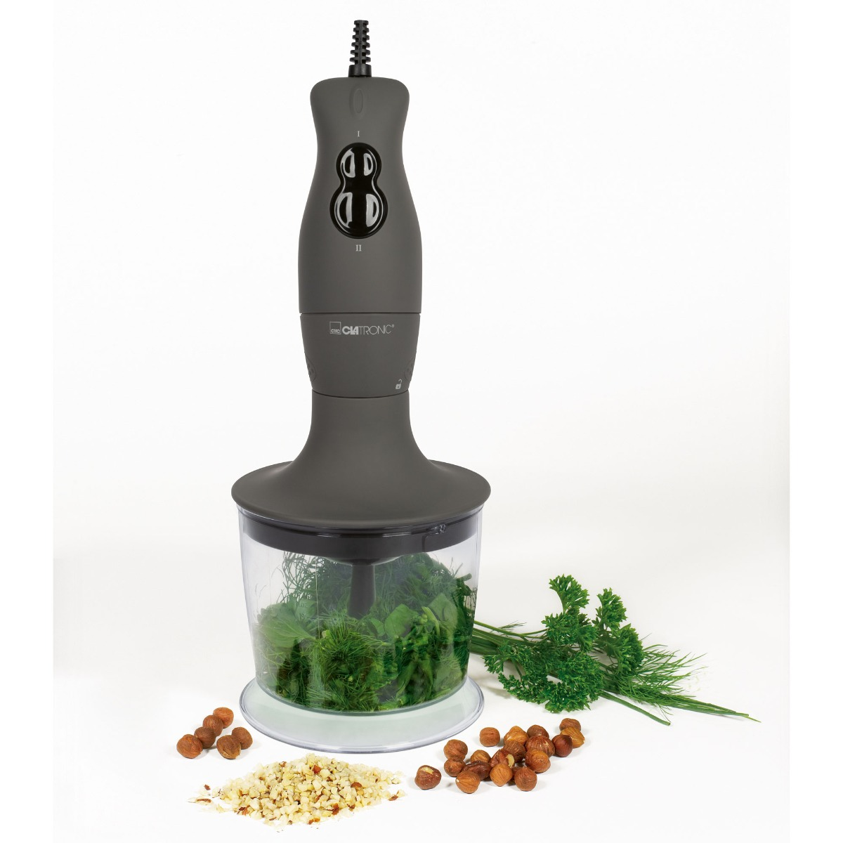 Szary blender ręczny z przezroczystym pojemnikiem wypełnionym zielonymi ziołami i orzechami laskowymi na białej powierzchni.