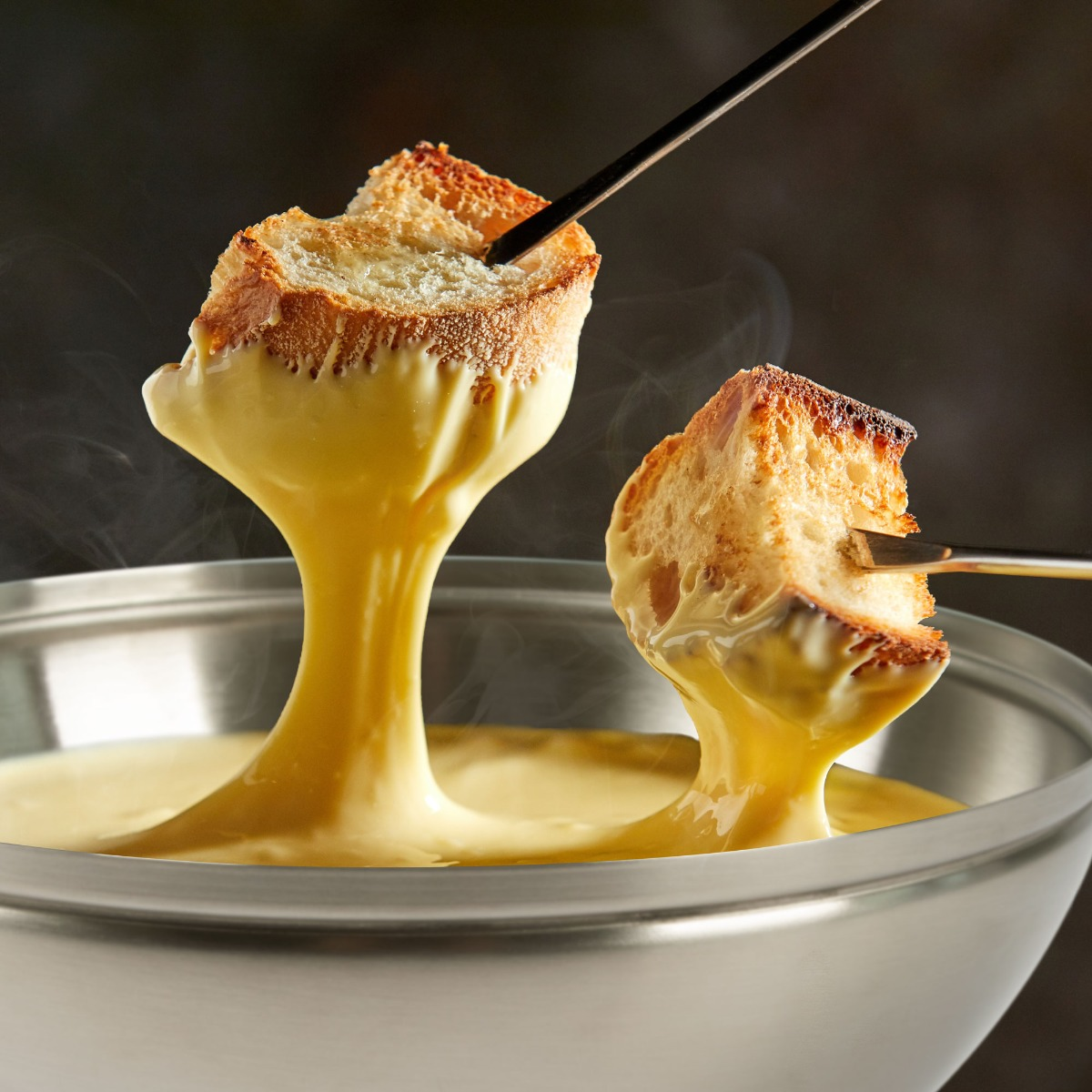 Nahaufnahme von Käsefondue mit Brotscheiben auf Gabeln, in einer Metallschüssel.
