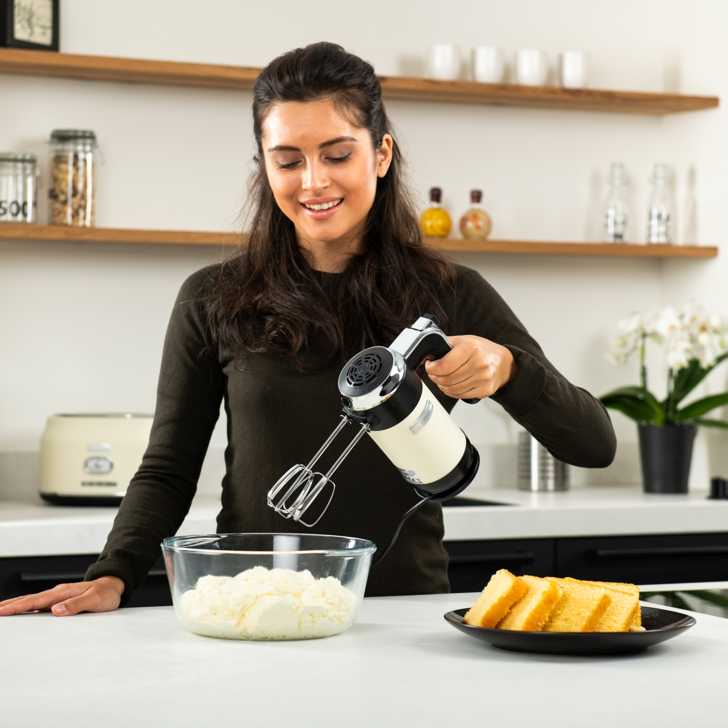 Frau, die mit einem Handrührgerät rührt. Auf der Theke stehen eine Glasschüssel und Kuchenstücke.