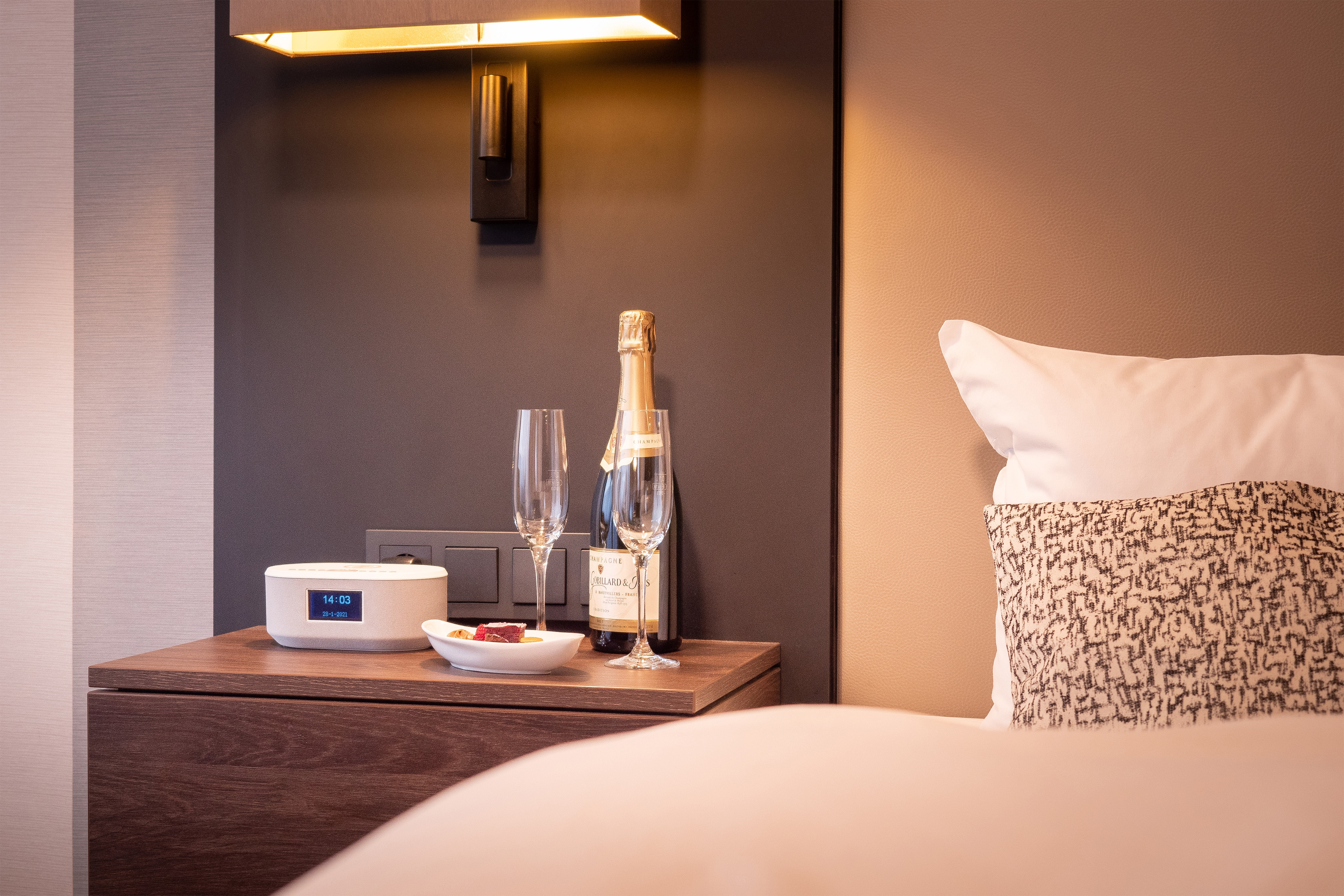 Nachttisch im Schlafzimmer mit Lampe, Champagner, Gläsern und kabellosem Lautsprecher. Ein Bett im Vordergrund.