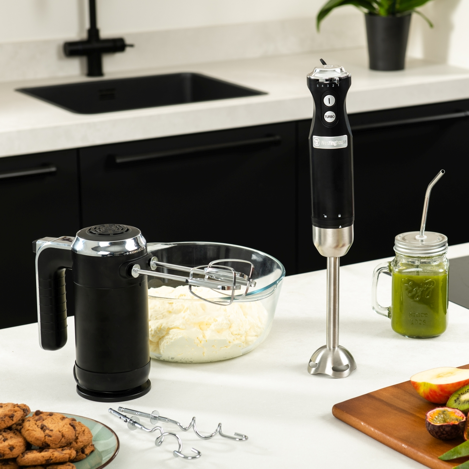 Handmixer und Stabmixer auf einer weißen Arbeitsplatte. Ein Smoothie und Kekse sind ebenfalls vorhanden.