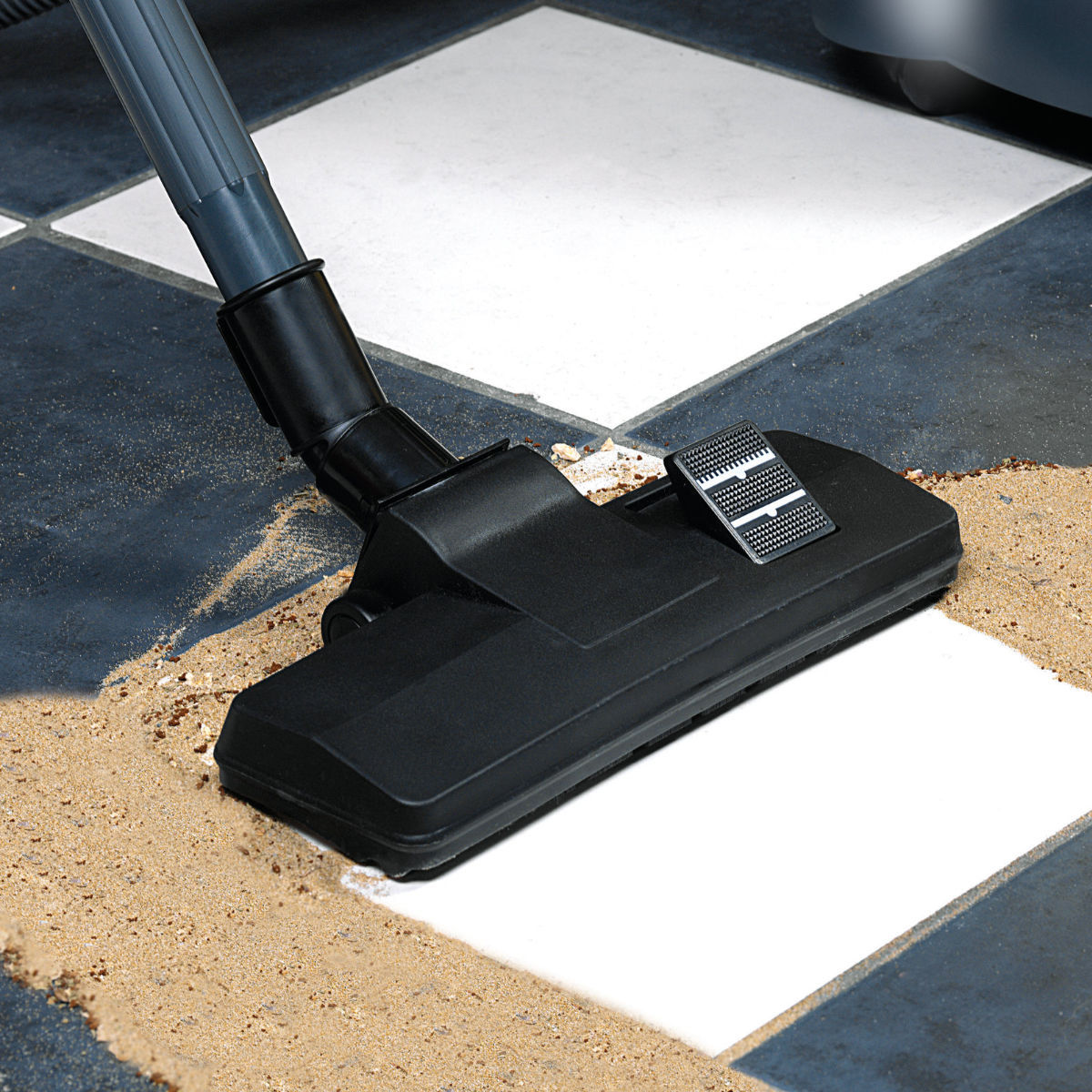 Schwarzer Staubsaugerkopf auf kariertem Boden, der Sand aufsaugt. Der Boden ist schwarz und weiß.