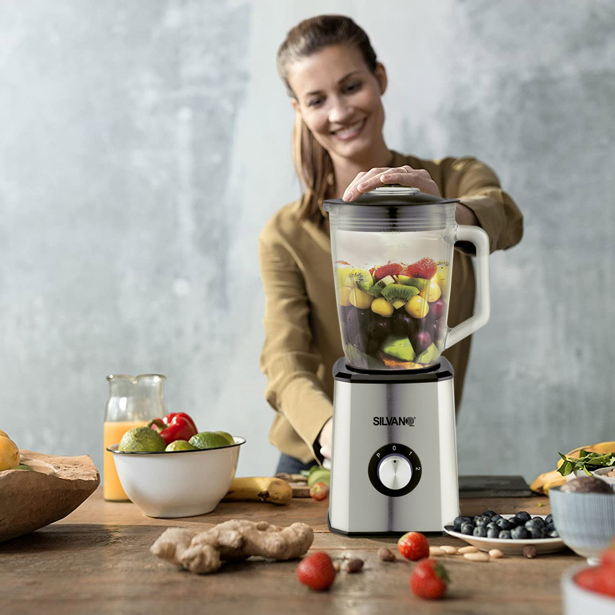 Mujer preparando un batido en una licuadora. Frutas en la mesa, incluyendo kiwi, arándanos y fresas.