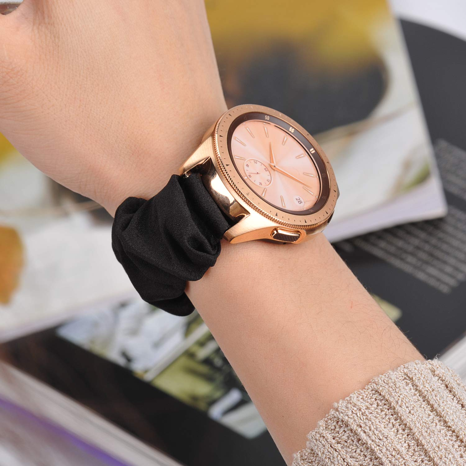 Roségoldene Smartwatch am Handgelenk mit schwarzem Scrunchie. Hintergrund: Bücher und ein beiger Pullover.
