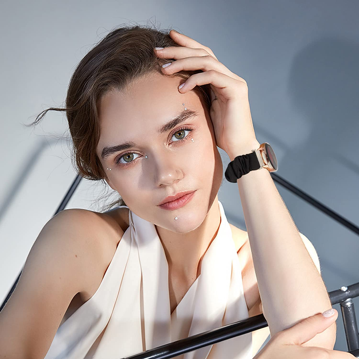 Eine Frau mit braunen Haaren trägt eine schwarze Uhr. Sie hat ein Piercing im Gesicht und lehnt sich an Metall.