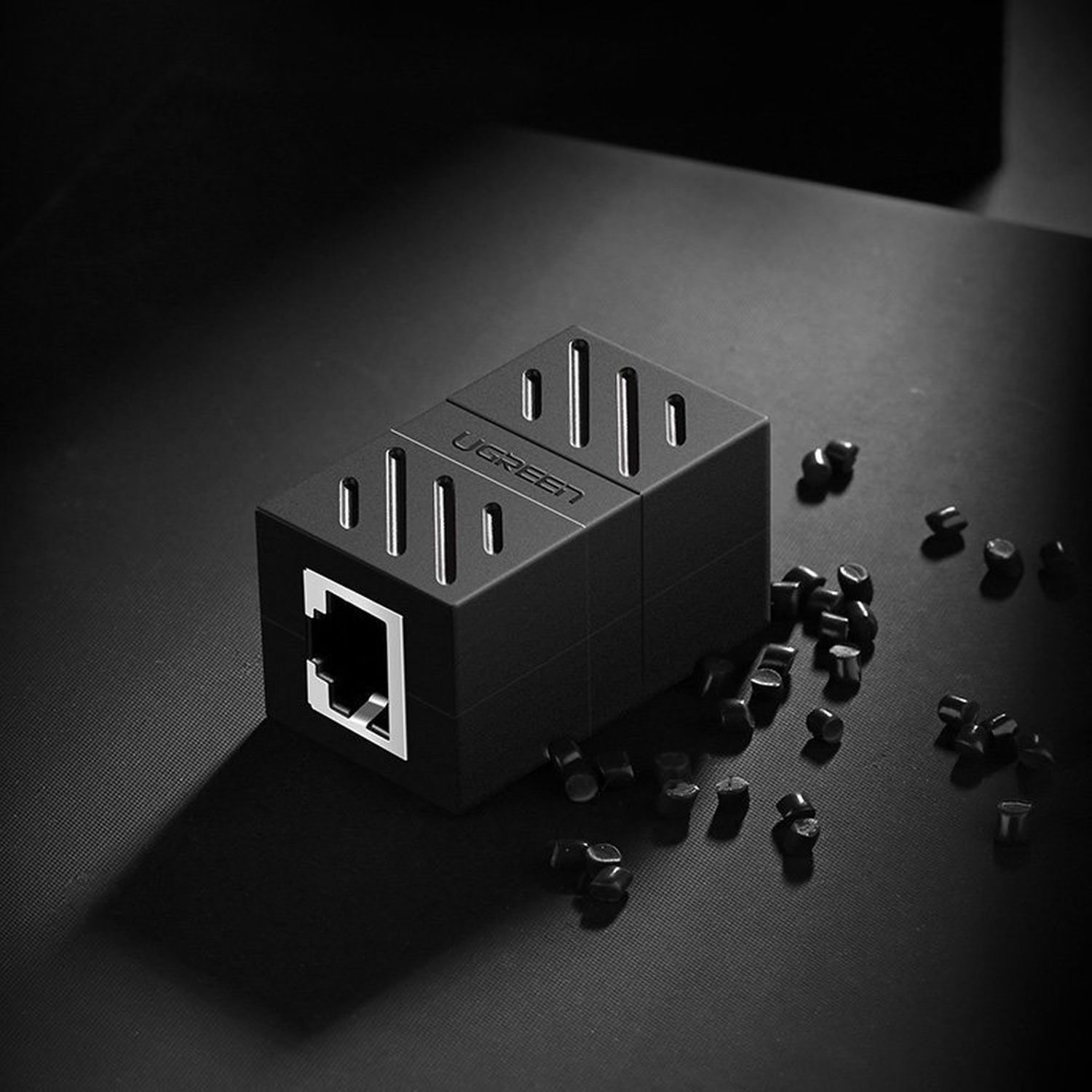 Ein schwarzer UGREEN-Netzwerkadapter in Würfelform mit einem Ethernet-Anschluss und verstreuten schwarzen Pellets.