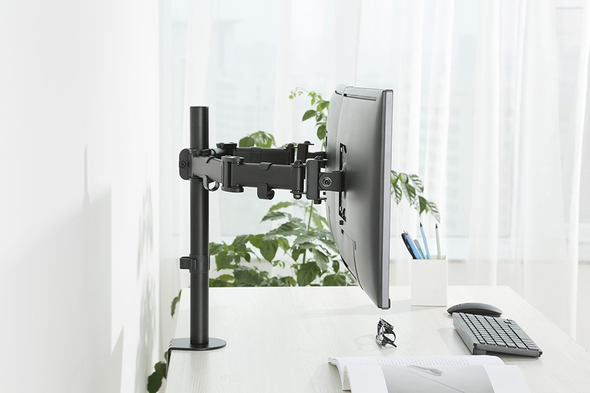 Monitorarm an einem Schreibtisch befestigt. Monitor ist sichtbar, zusammen mit Tastatur, Maus und Pflanze.