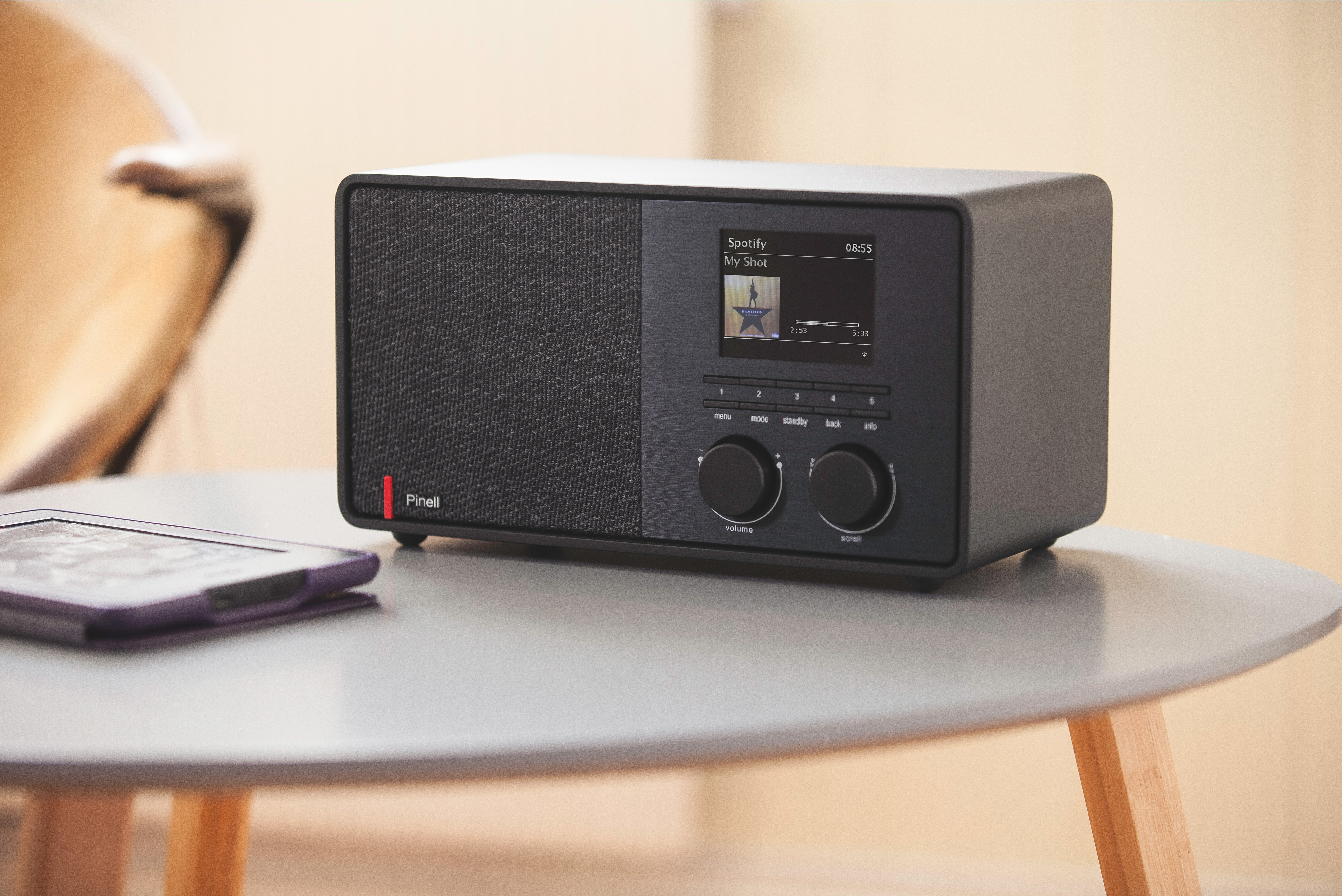 Une radio noire posée sur une table, affichant une liste de lecture Spotify, avec un petit lecteur ebook à côté.