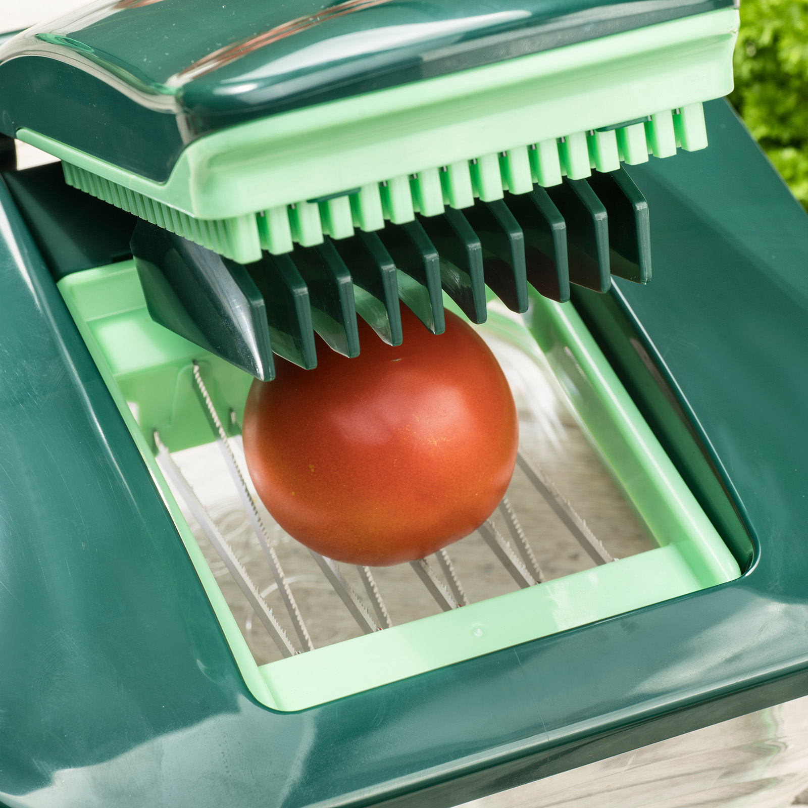 Eine Tomate wird von einem grünen Küchenwerkzeug geschnitten. Das Werkzeug hat Klingen und einen grünen Rahmen.